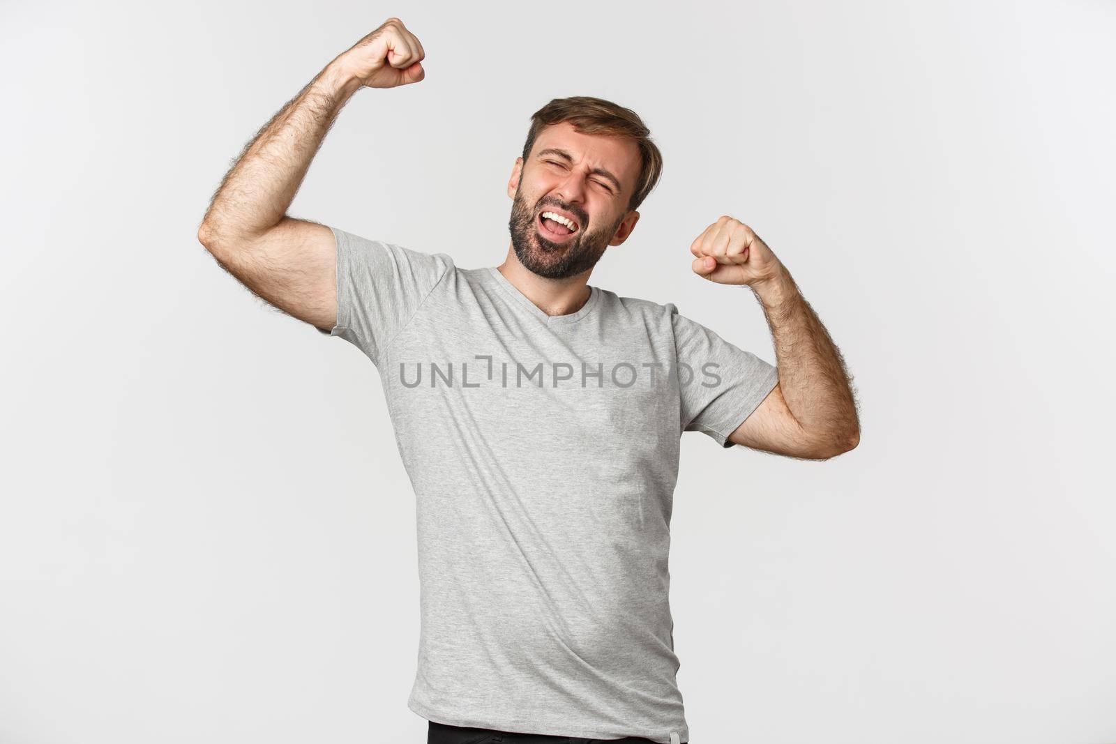 Portrait of excited and happy man in gray t-shirt, making fist pump and shouting yes to celebrating win, triumphing over white background.