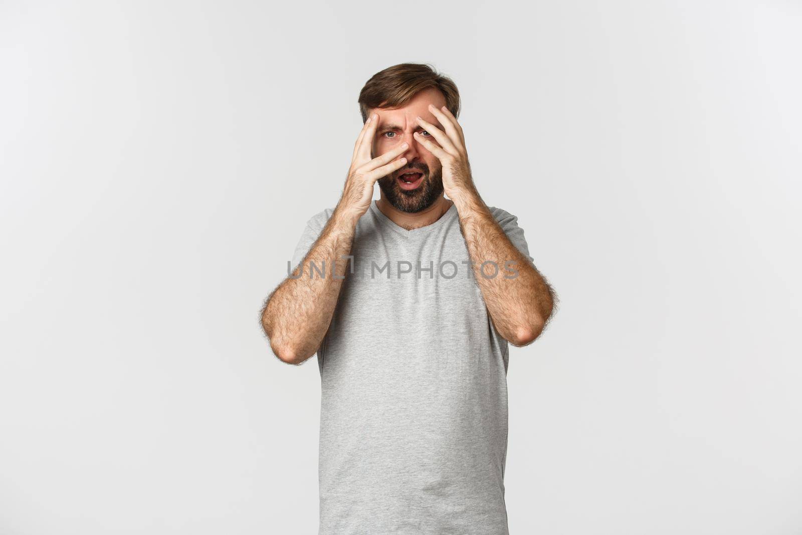 Portrait of embarrassed bearded man in gray t-shirt, cover eyes with hands but peeking through fingers scared to look, standing over white background by Benzoix