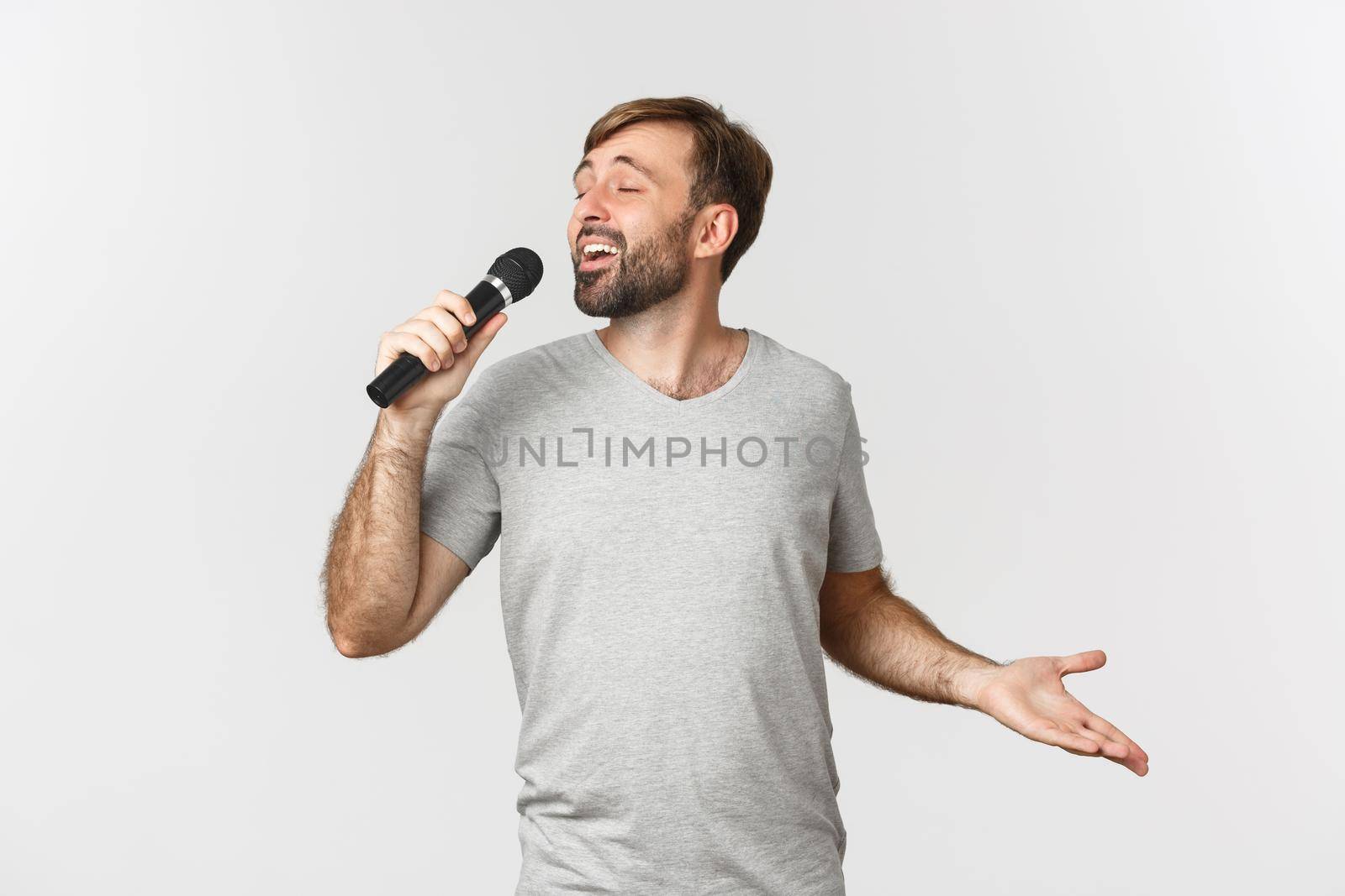 Handsome carefree guy singing song in karaoke, holding microphone, standing over white background.