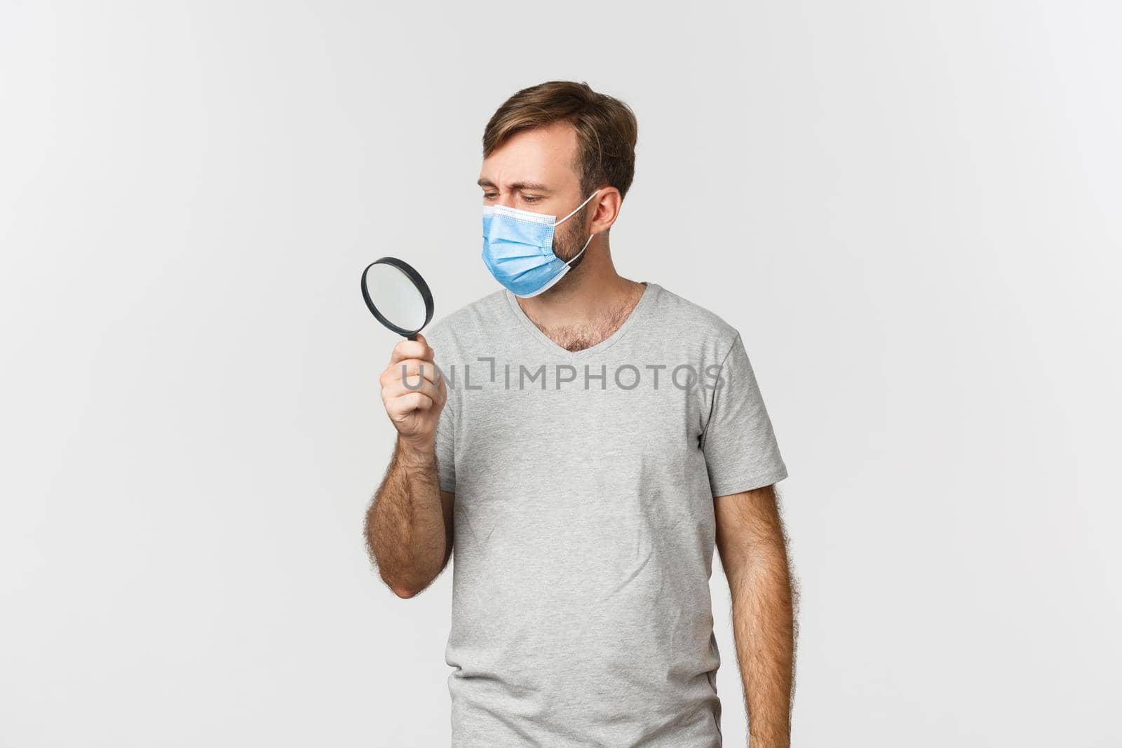 Concept of pandemic, covid-19 and social-distancing. Image of serious man in medical mask, searching for something and looking through magnifying glass, standing over white background.