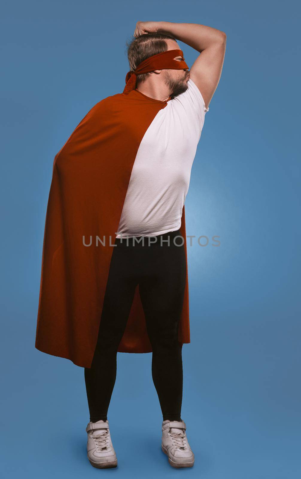 Super hero man ready to save world. Serious man shows and kisses his muscles standing in red super hero mask and coat. Cut out on faded denim blue background.