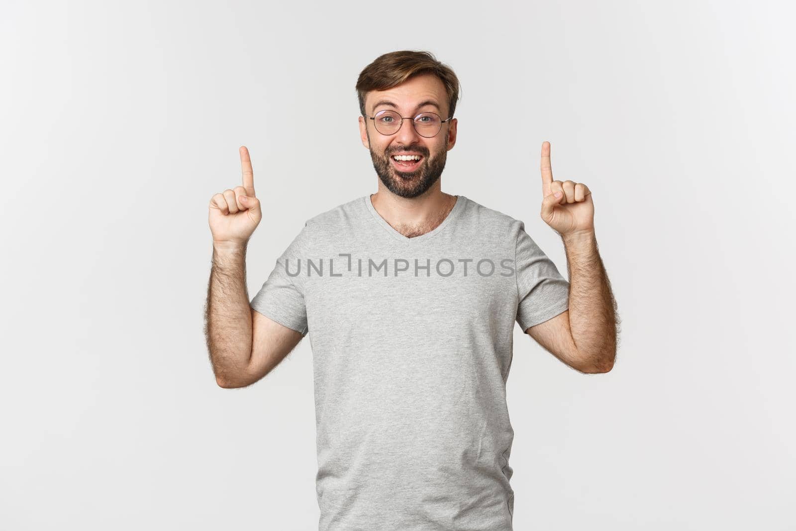 Cheerful bearded man smiling, pointing fingers up, showing logo, wearing gray t-shirt, standing over white background.