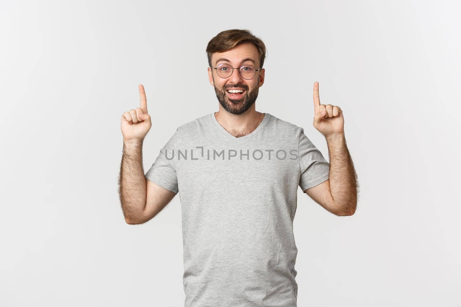 Cheerful bearded man smiling, pointing fingers up, showing logo, wearing gray t-shirt, standing over white background by Benzoix