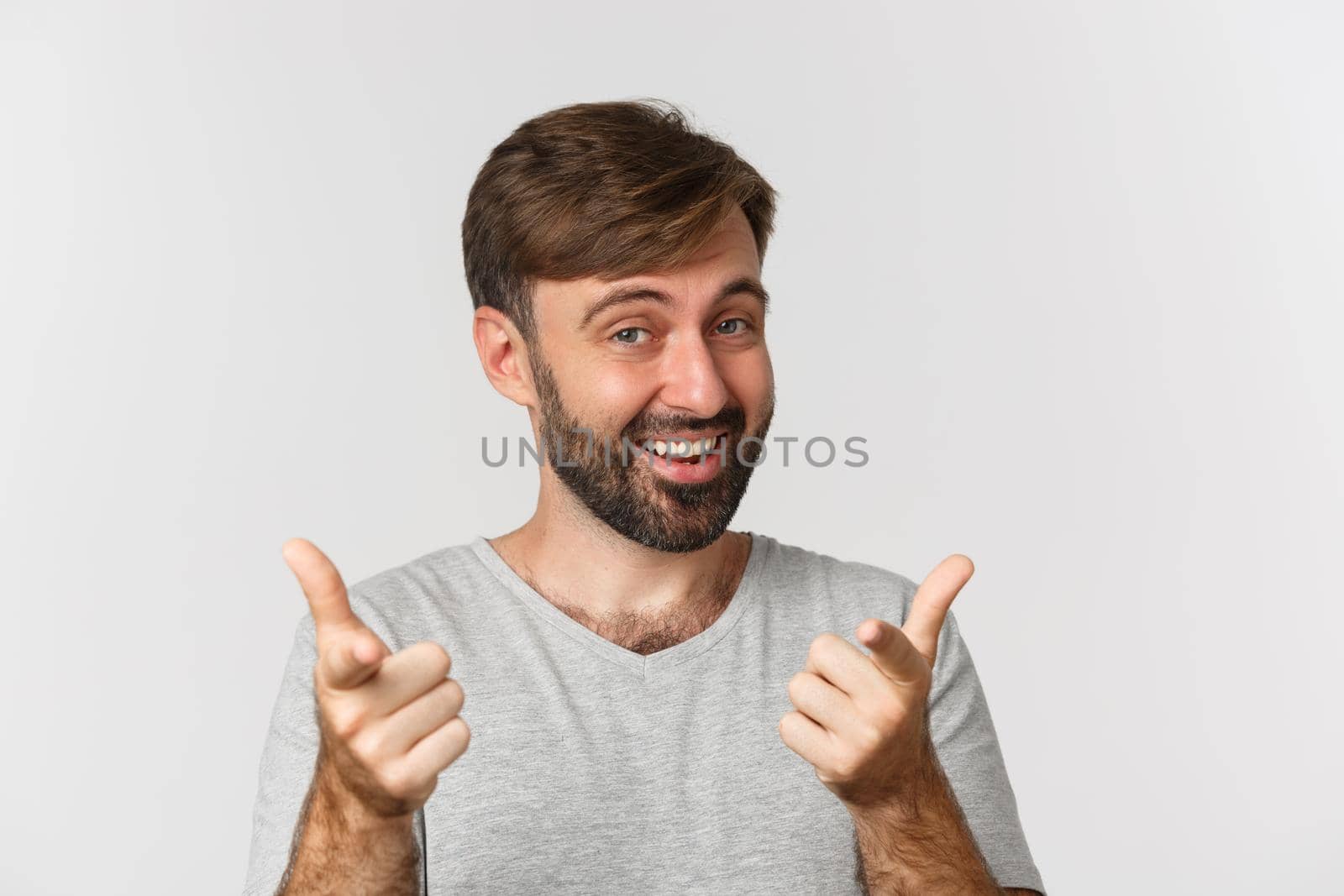 Close-up of handsome adult man with beard, smiling and pointing fingers at camera, standing in gray t-shirt over white background.