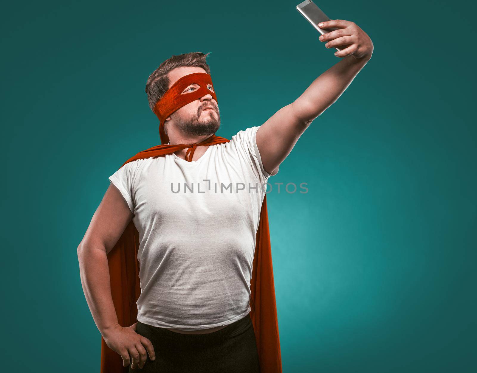 Super Hero man Takes Selfie By Mobile Phones. Man In A Super Hero Costume In A Red Mask And Cloak Isolated On Biscay Green Background.