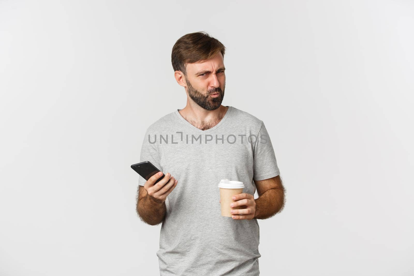 Image of confused bearded man standing with coffee and mobile phone, looking puzzled.
