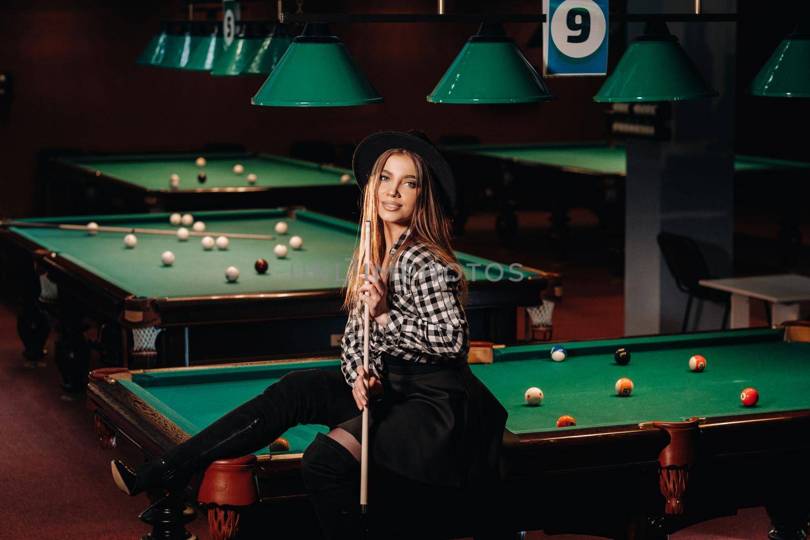 A girl in a hat in a billiard club sits on a billiard table with a cue in her hands.Playing billiards by Lobachad