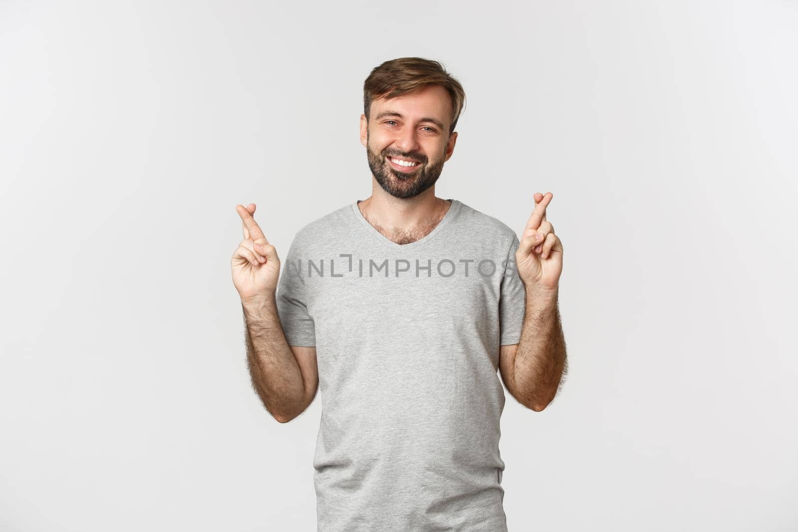 Portrait of optimistic handsome man in gray t-shirt, hoping for something with fingers crossed, making wish and smiling, standing over white background.