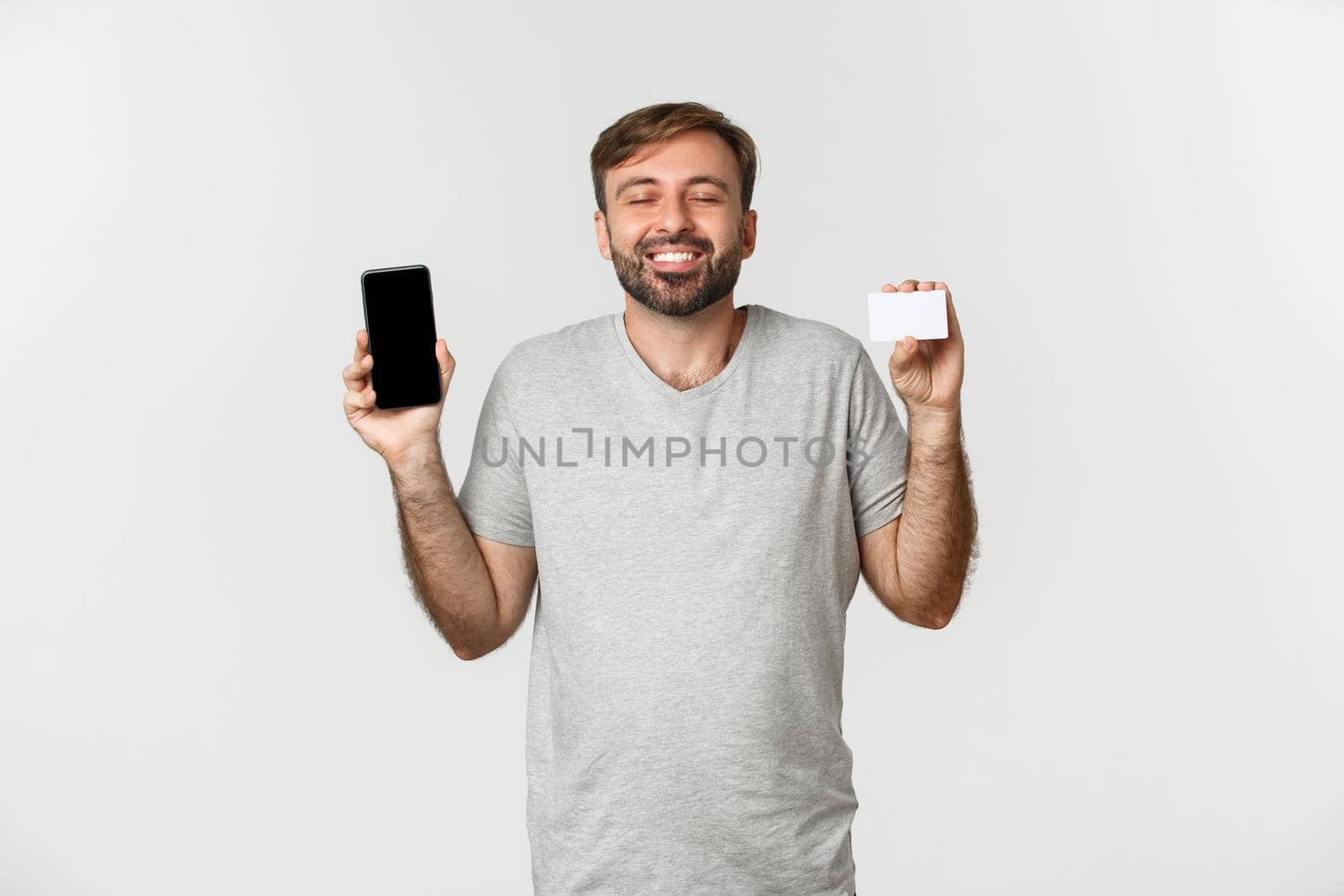 Handsome young man shopping online, holding credit card and mobile phone, showing smartphone screen, standing over white background.