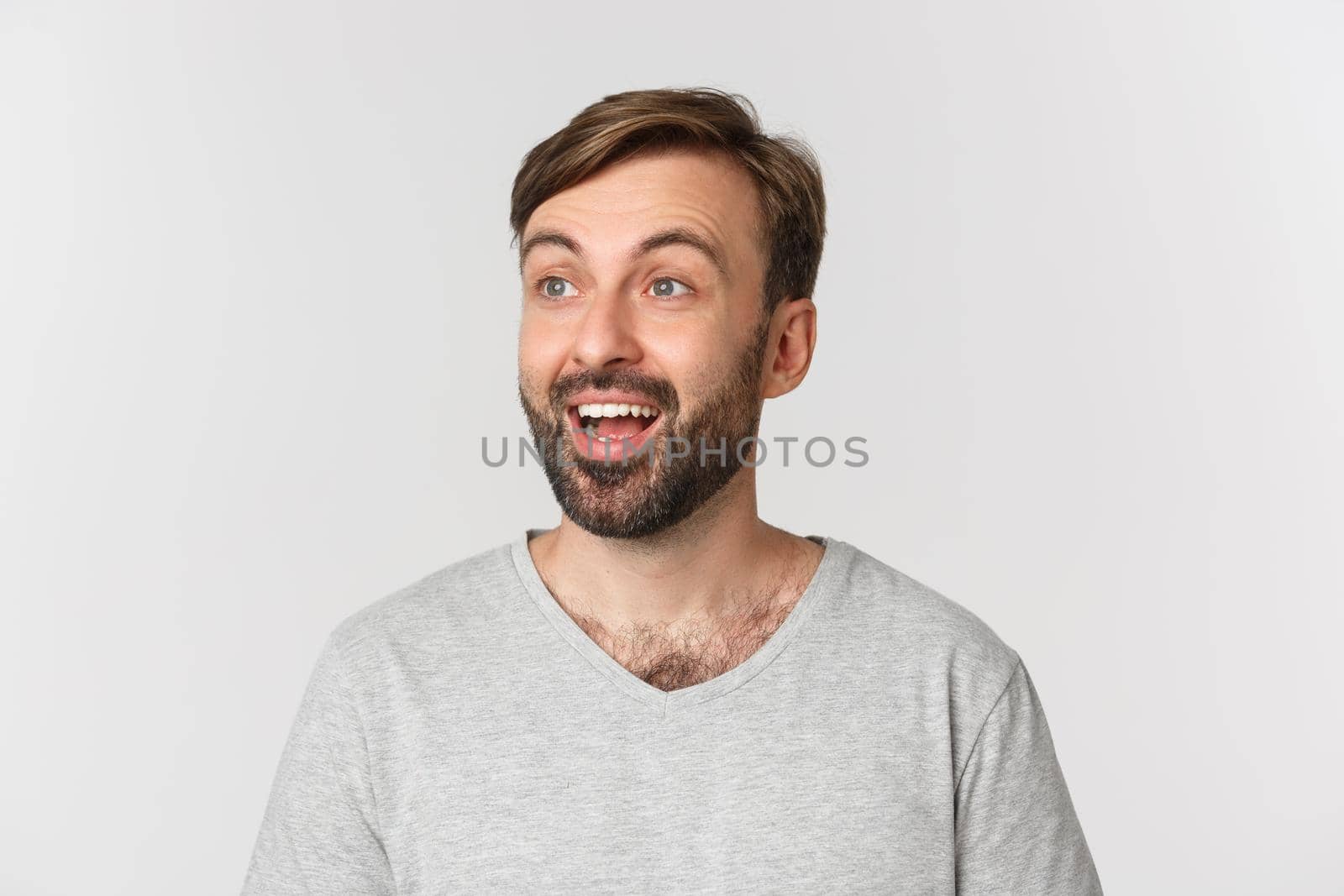 Portrait of handsome bearded man in gray t-shirt, looking left with happy and excited face, standing over white background.