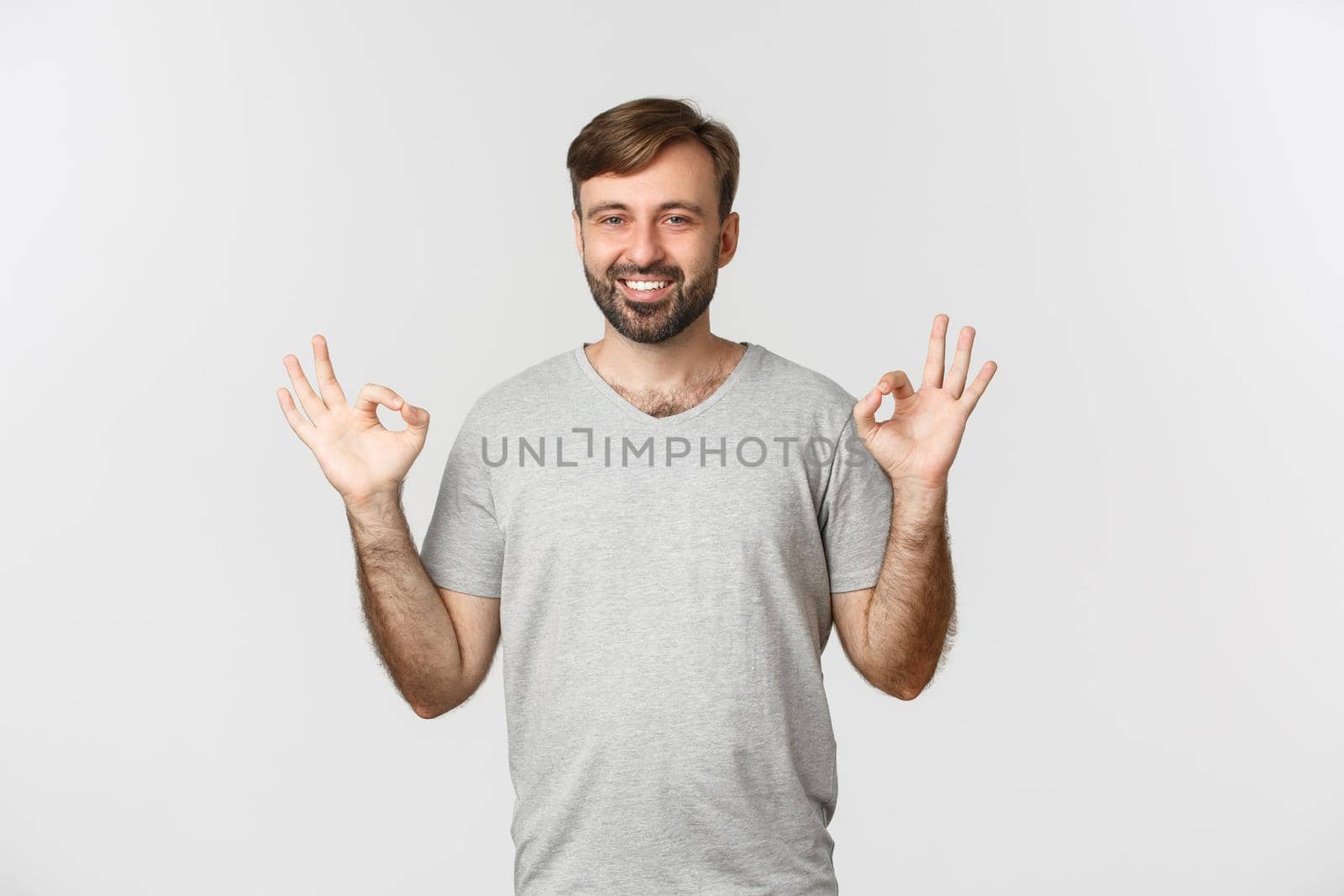 Portrait of handsome smiling man in casual grey t-shirt, showing okay sign in approval, agree to you, praising good choice, standing over white background by Benzoix