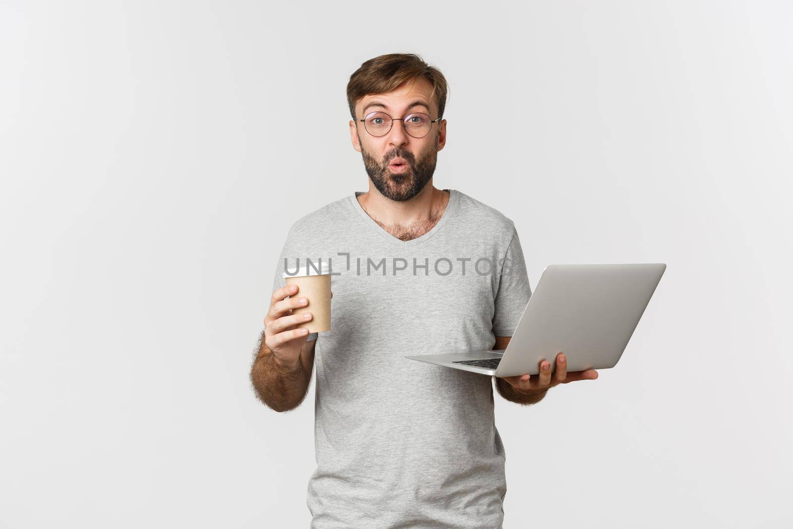 Portrait of handsome male freelancer, drinking coffee and working with laptop, looking surprised, standing over white background.
