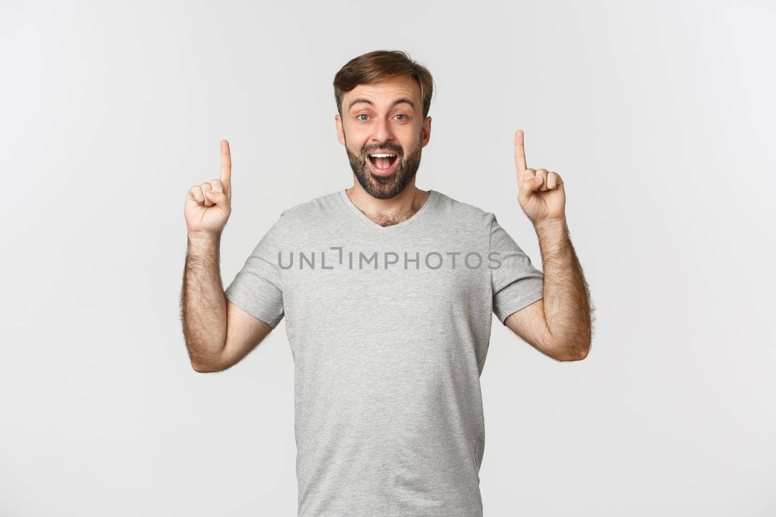 Cheerful handsome man in gray t-shirt, showing logo or advertisement, pointing fingers up and smiling happy, standing over white background by Benzoix