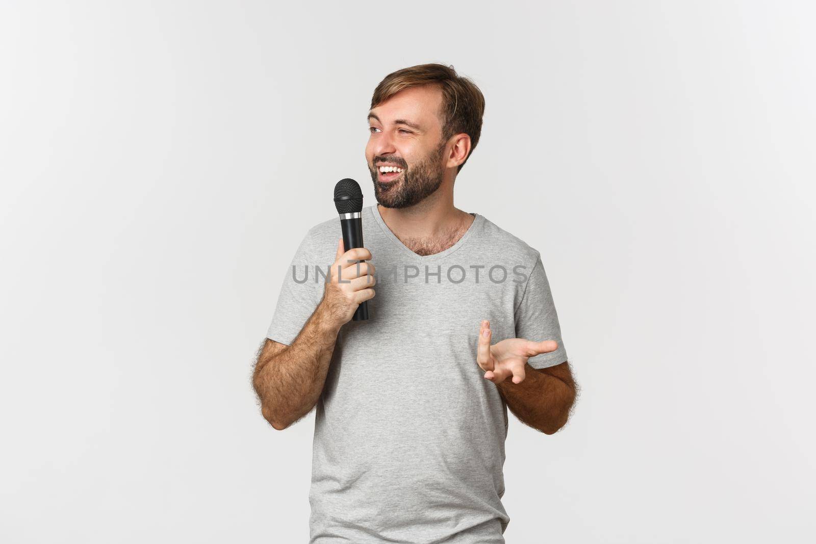 Image of charismatic male comedian in gray t-shirt making a speech, holding microphone and talking, standing over white background.