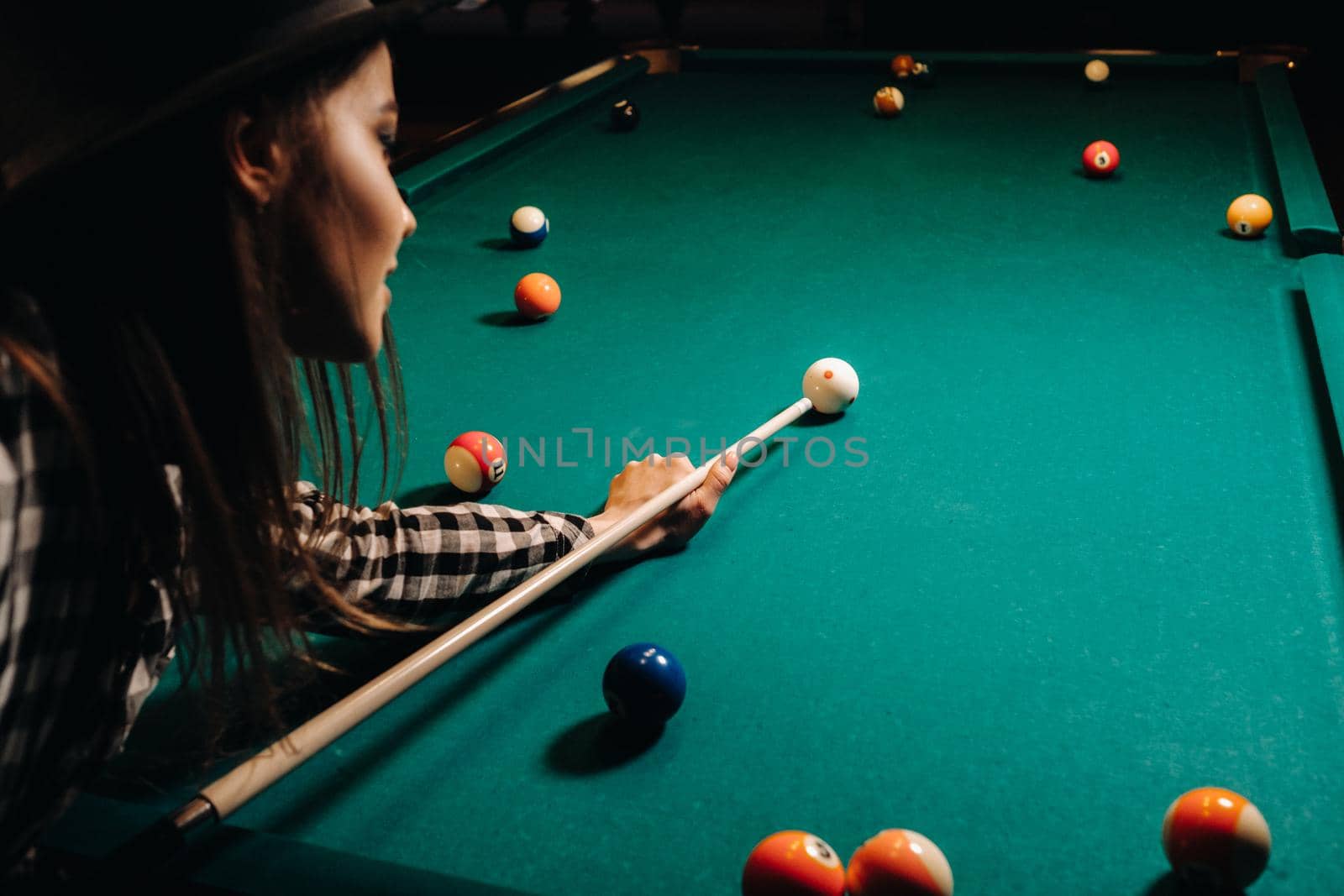 A girl in a hat in a billiard club with a cue in her hands hits a ball.Playing billiards.