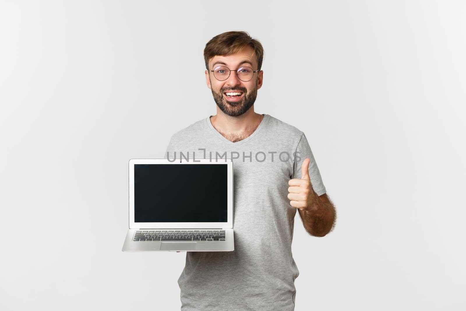 Cheerful man in gray t-shirt, showing laptop screen and smiling, making thumbs-up in approval, standing over white background.