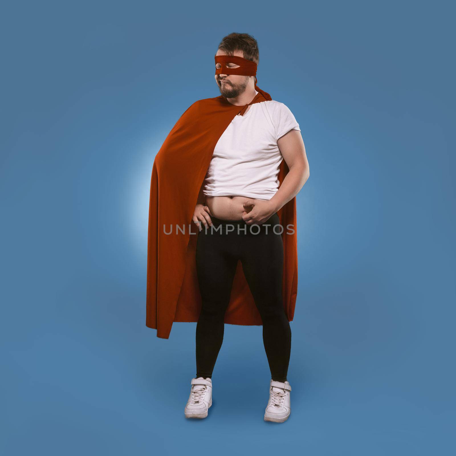 Body positive Super hero man added weight over winter. Sad man in superhero costume checks fat on his tummy. Isolated on blue background. Template with place for text. Overweight concept by LipikStockMedia