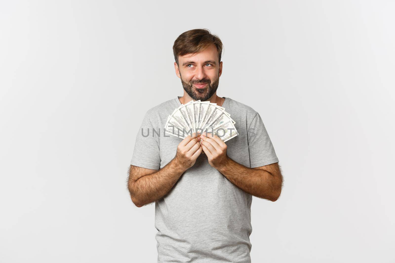 Greedy smiling man with beard, thinking about shopping, holding money and looking at upper left corner, standing over white background.