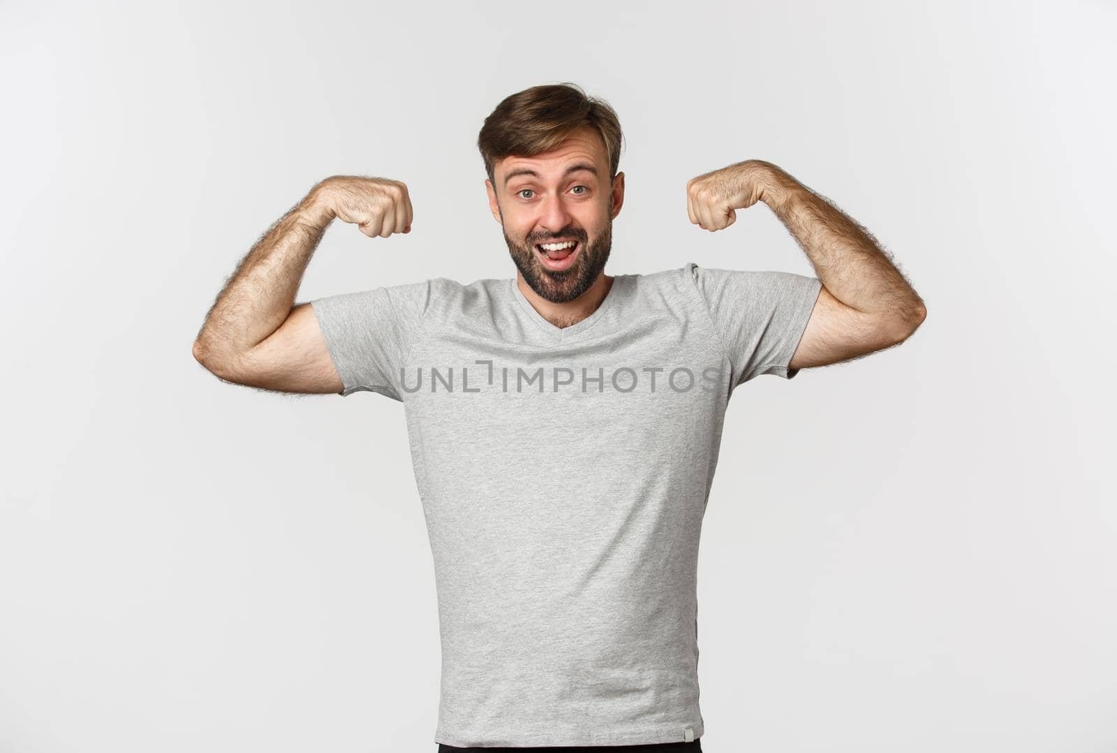 Portrait of confident bearded man flexing biceps, showing his muscles after gym workout, standing over white background.