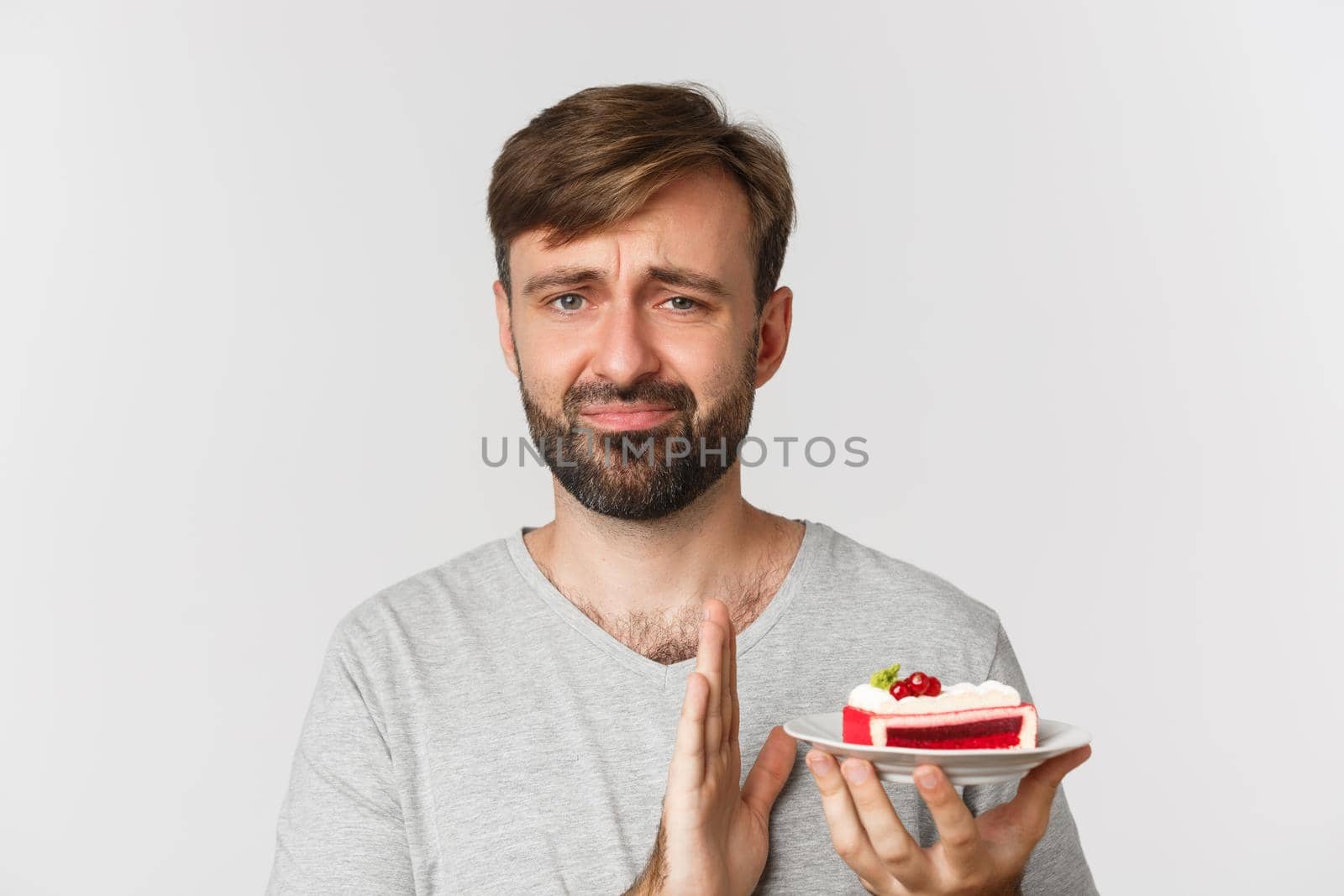 Close-up of reluctant bearded man, declining piece of cake and grimacing, standing over white background by Benzoix