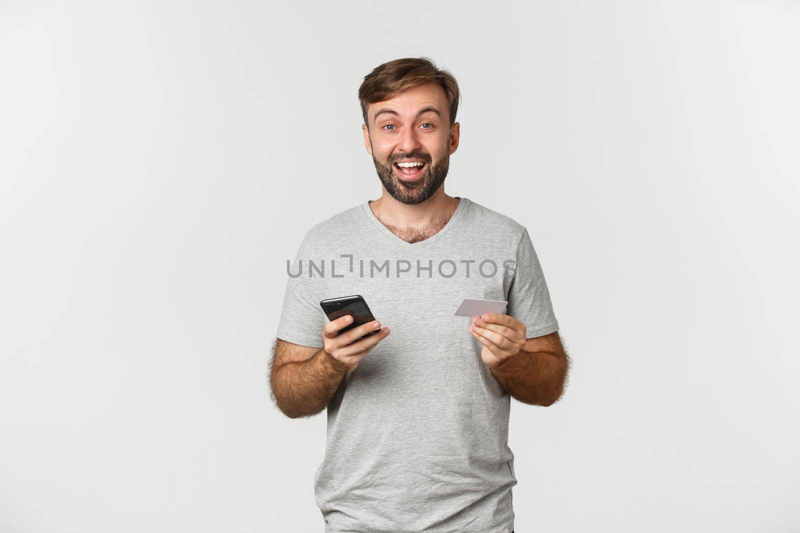 Handsome young man shopping online, holding credit card and mobile phone, standing over white background.