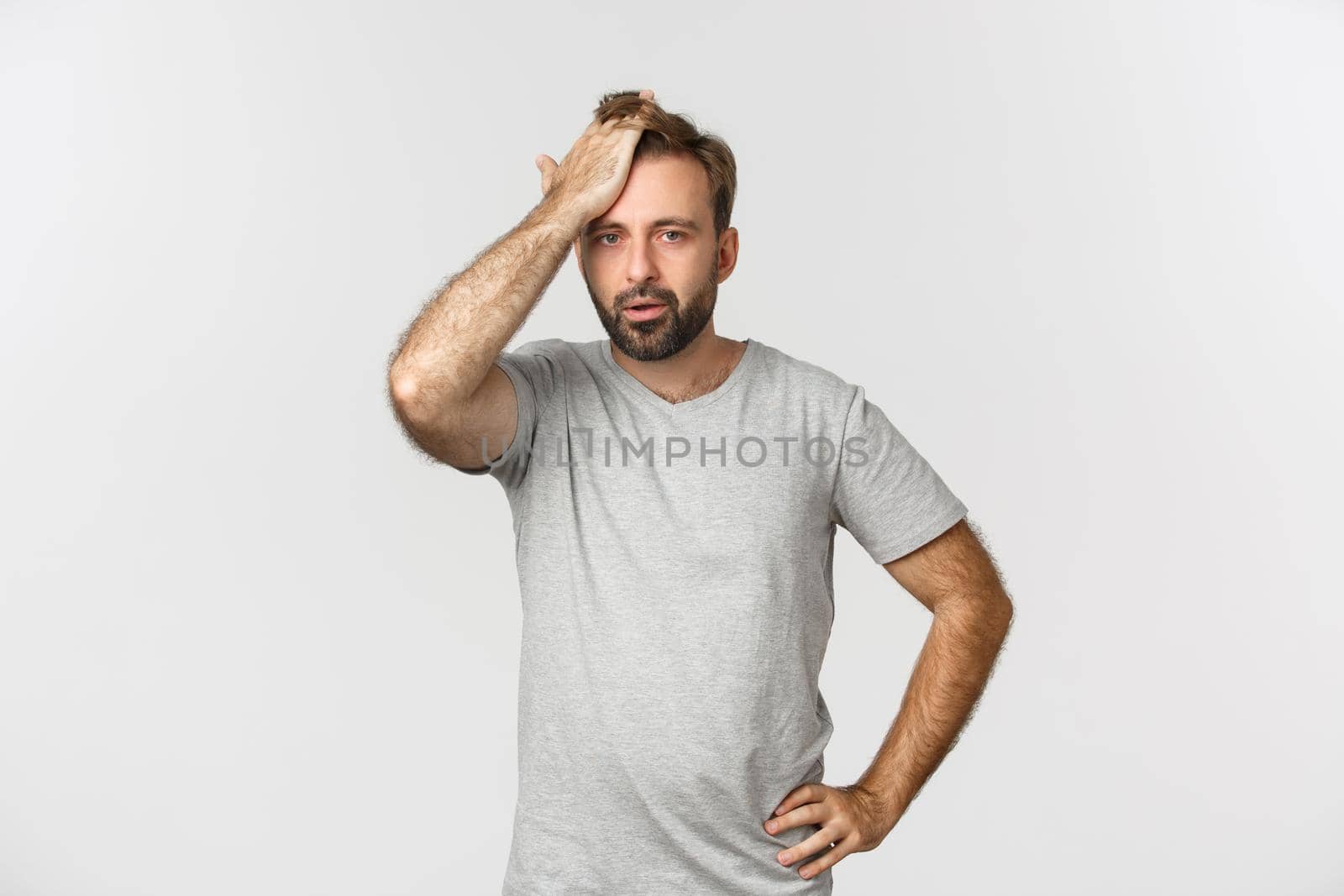 Troubled bearded man in gray t-shirt, slap forehead and sighing bothered by problem, standing over white background distressed.