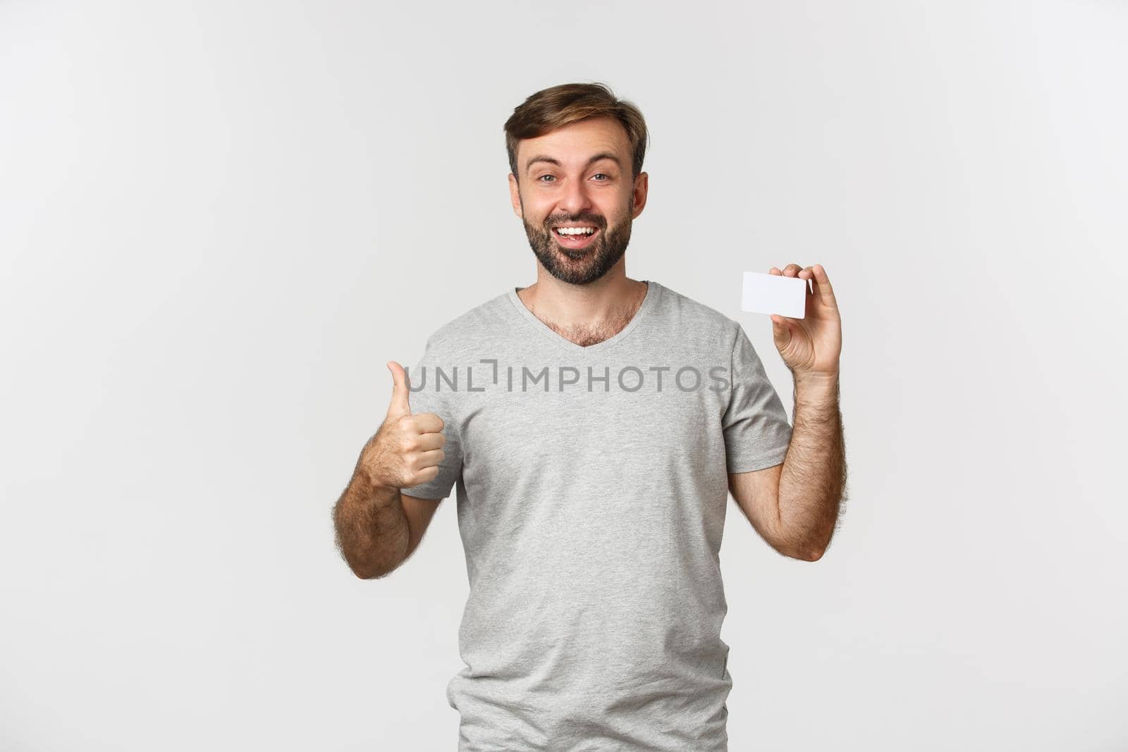 Portrait of handsome smiling man in gray t-shirt, showing credit card, making thumbs-up in approval, standing over white background.