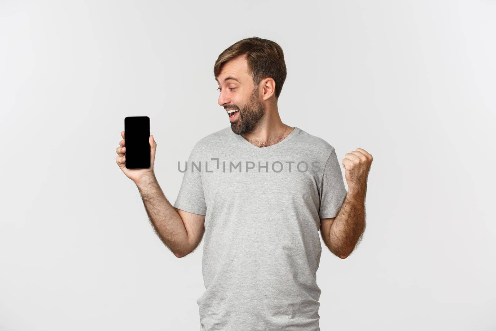 Portrait of excited man with beard, achieve goal in app, showing mobile phone screen and rejoicing, winning something, standing over white background.