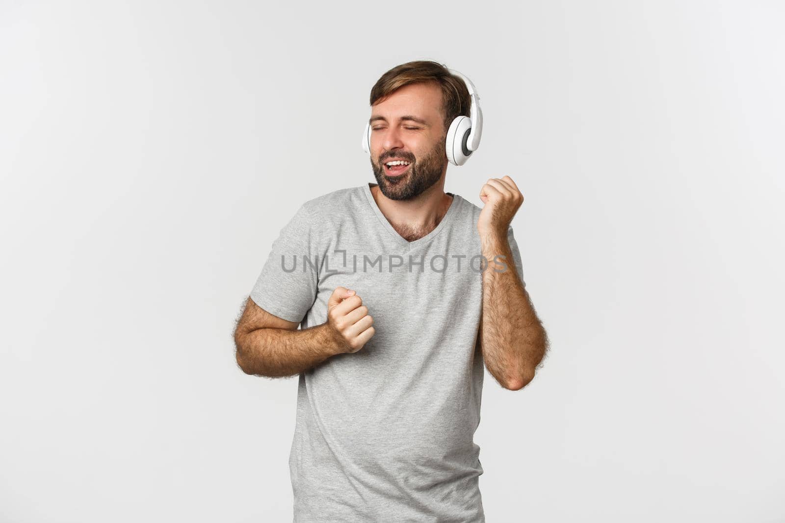 Portrait of happy handsome guy in gray t-shirt, listening music in wireless headphones and dancing, standing over white background by Benzoix