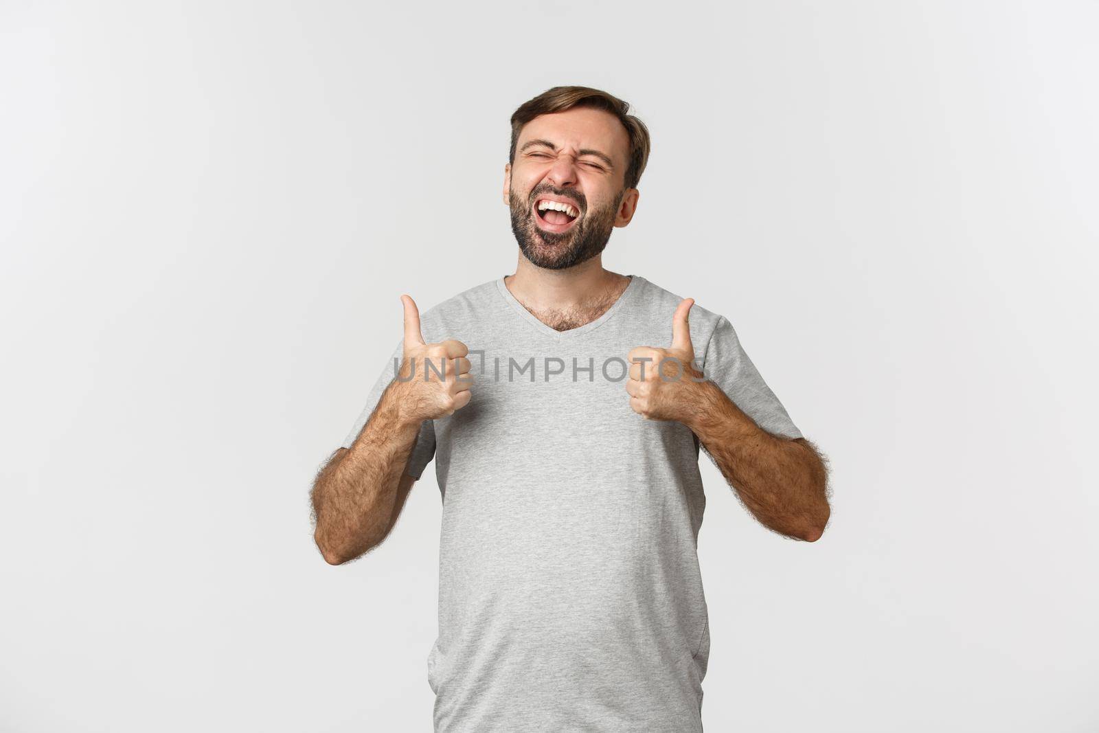 Portrait of happy and excited guy with beard, laughing over something funny and showing thumbs-up, standing over white background.