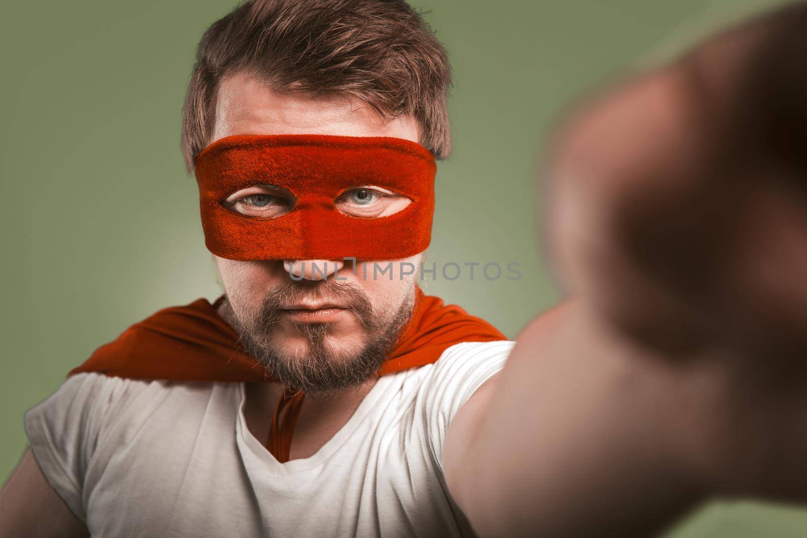 Super hero man makes selfie photos or video by mobile phones. Serious man wearing red mask and cloak posing for his social networks followers. Isolated on green background by LipikStockMedia