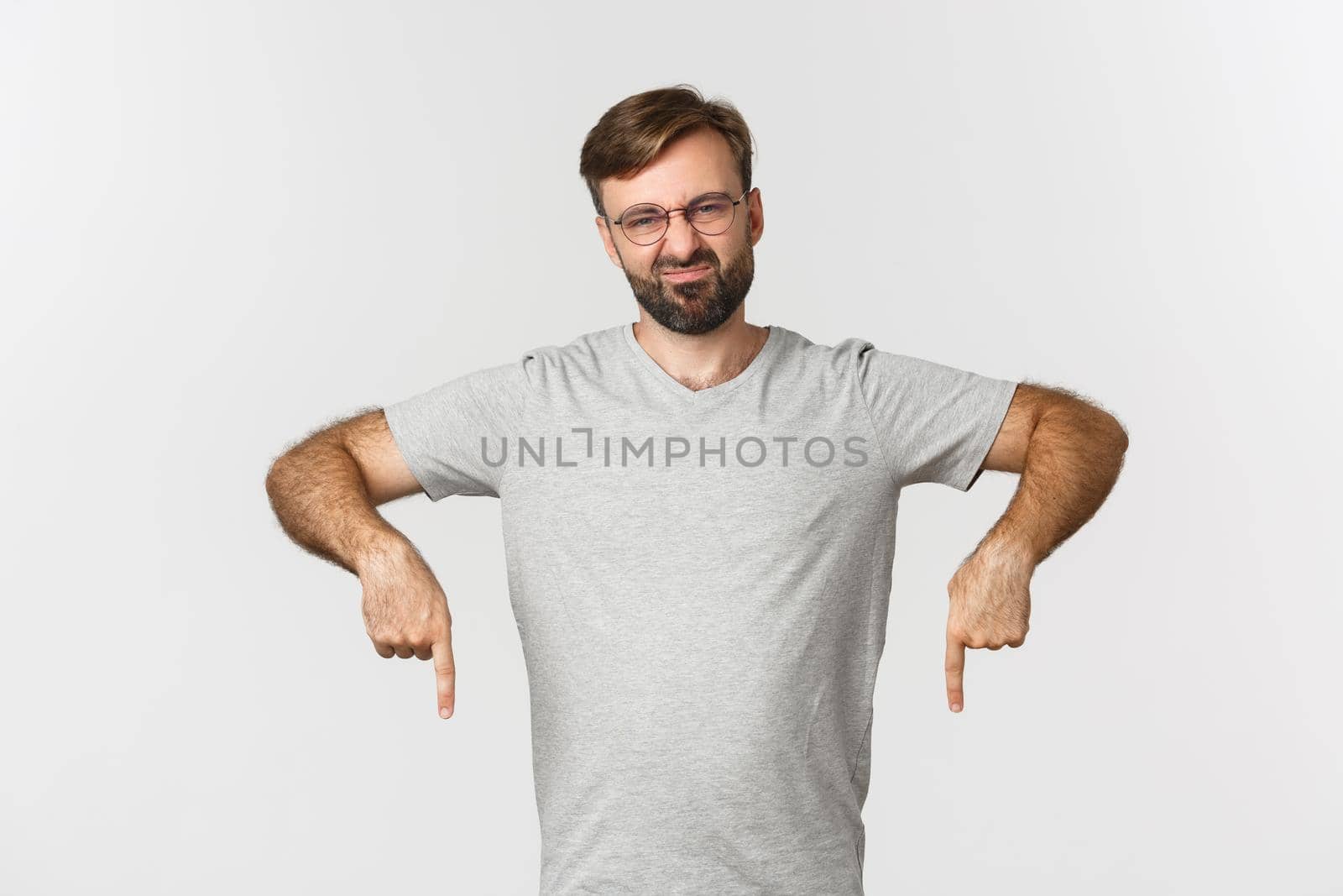 Disappointed bearded man grimacing, pointing fingers down, showing logo, wearing gray t-shirt, wearing gray t-shirt, standing over white background.