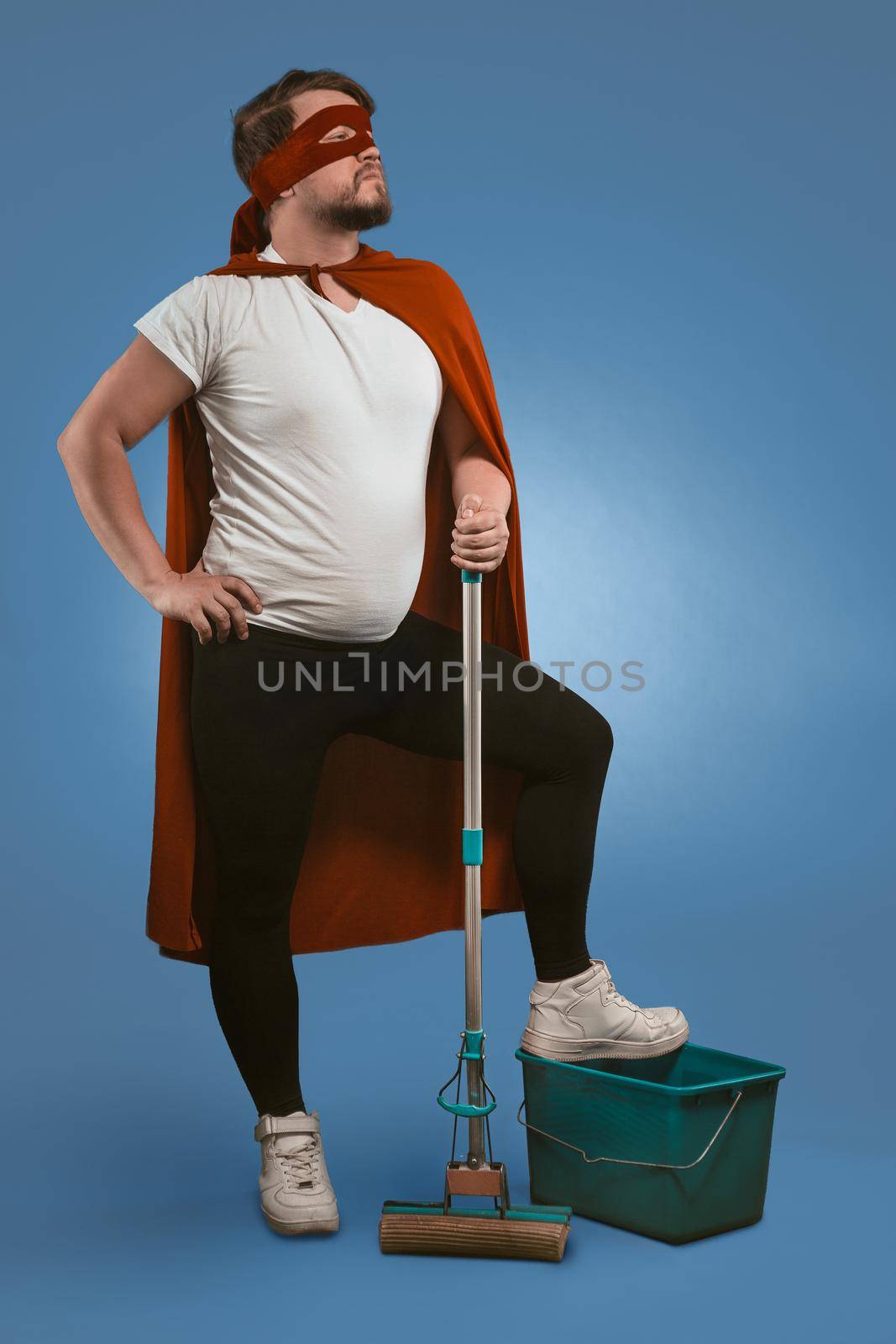 Super cleaning from superhero. Brave guy ready to clean up and fight dark forces with help of cleaning equipment. Caucasian man looks forward holding mop with his foot on bucket on blue background by LipikStockMedia