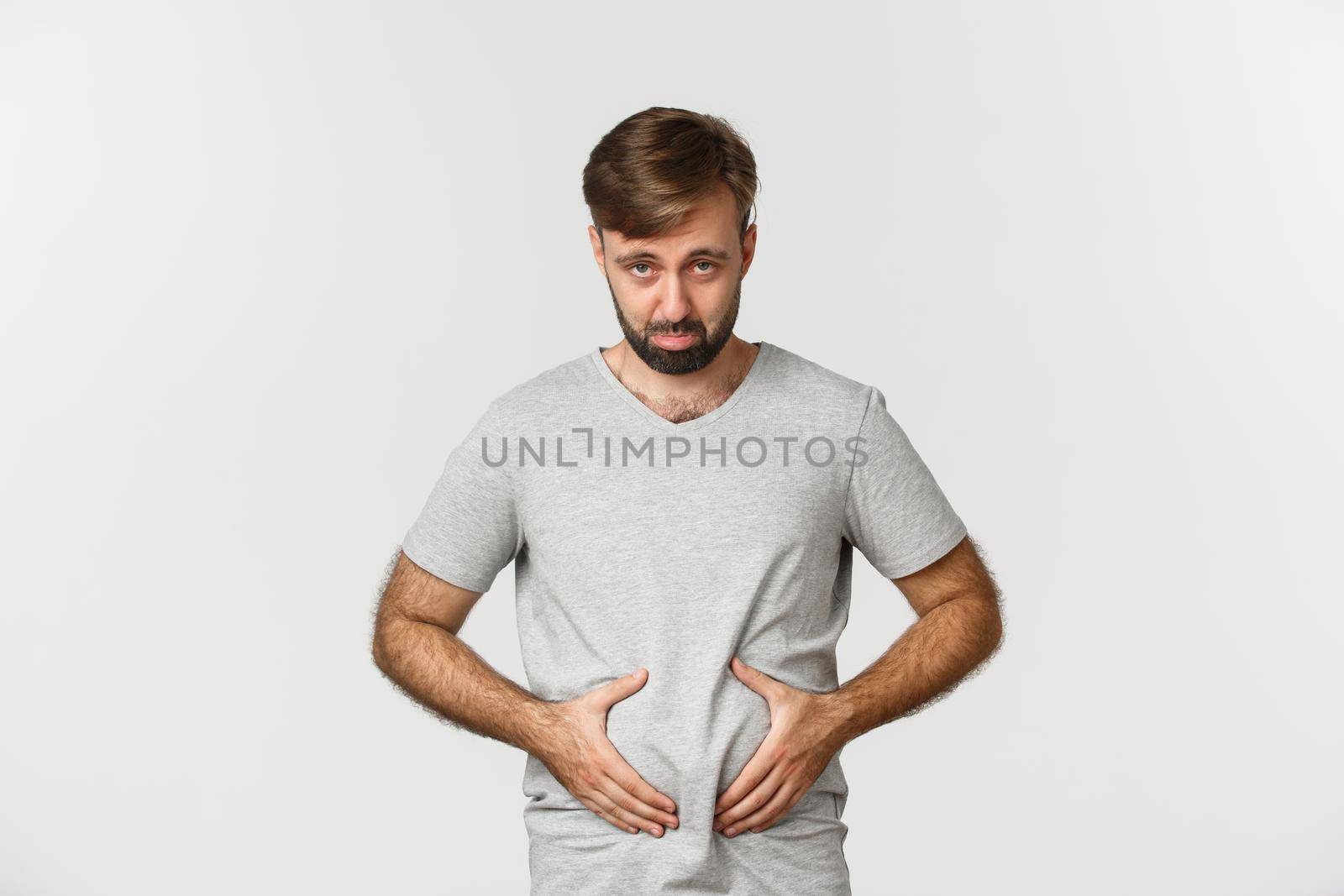 Portrait of guy feeling upset about his weight, showing fat on belly and looking disappointed, sulking as standing over white background.