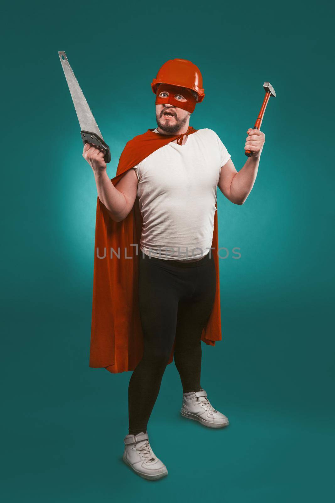 Super hero handyman holding saw and hammer. Man wearing red helmet, mask and cape holds building equipment standing on biscay green background. Handyman concept.