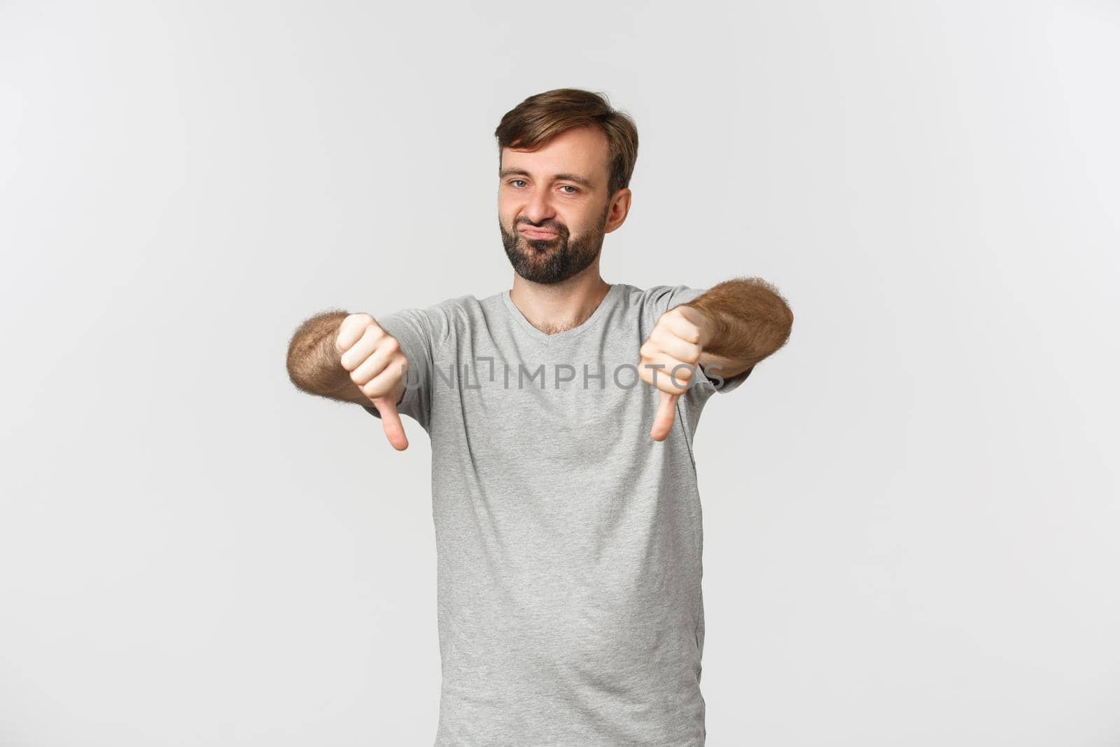 Portrait of skeptical bearded man in casual t-shirt, showing thumbs-down and looking disappointed, disapprove something bad, standing over white background by Benzoix