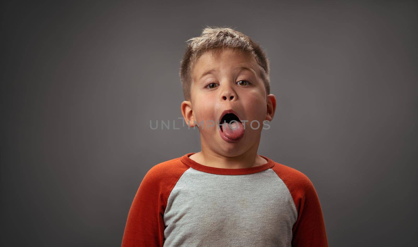 Preschool boy shows his tongue to camera. Portrait of funny kid on gray background. Emotions concept.