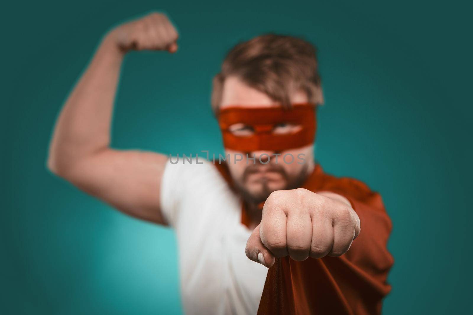 Brave Superhero man demonstrates muscles looking at camera. Attractive Caucasian guy in red mask and cloak ready to save world. Focus on fist in foreground. Cut out on biscay green background by LipikStockMedia