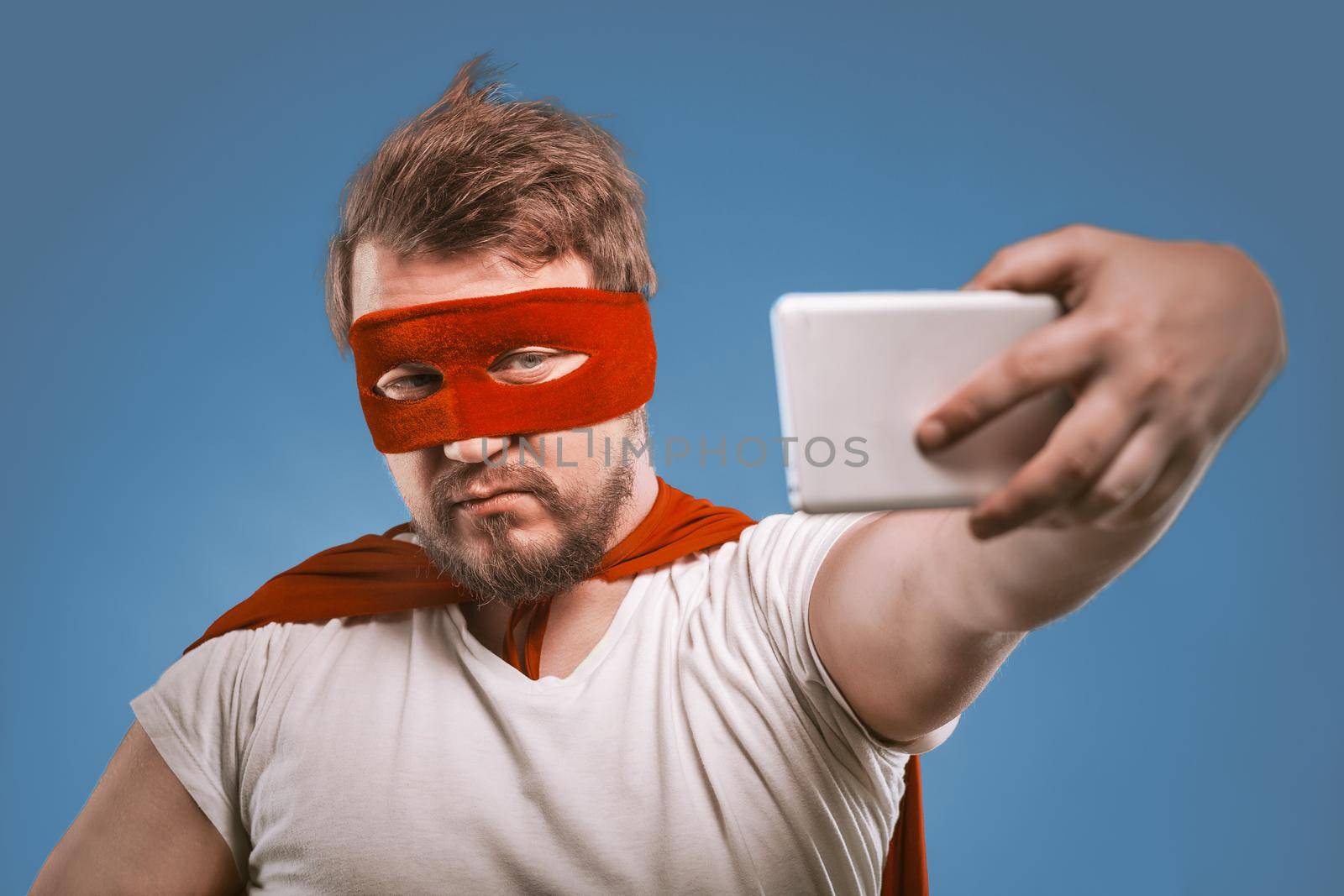 Super hero man takes selfie by mobile phones. Confident man in a super hero costume in a red mask and cloak poses on faded denim blue background making selfie photos on smartphone.