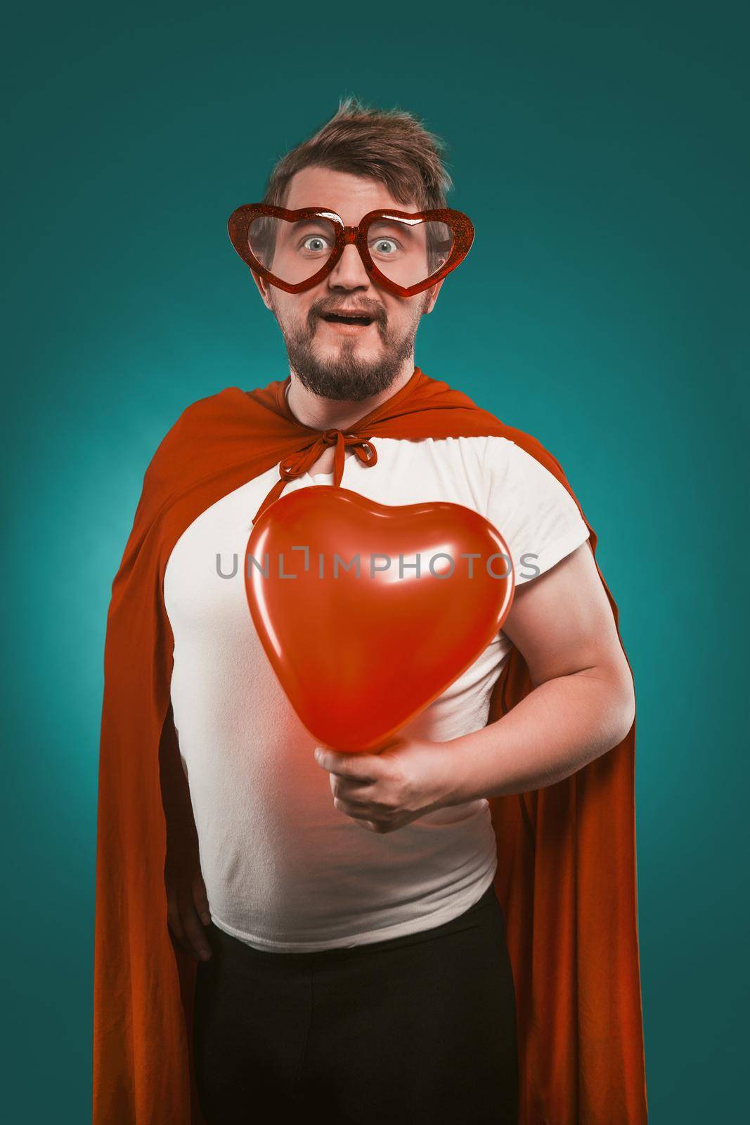 Superman In Love Holds Big Red Heart. Positive Man In Superhero Costume And Heart-Shaped Glasses Poses On Biscay Green Background. Valentine's day concept by LipikStockMedia