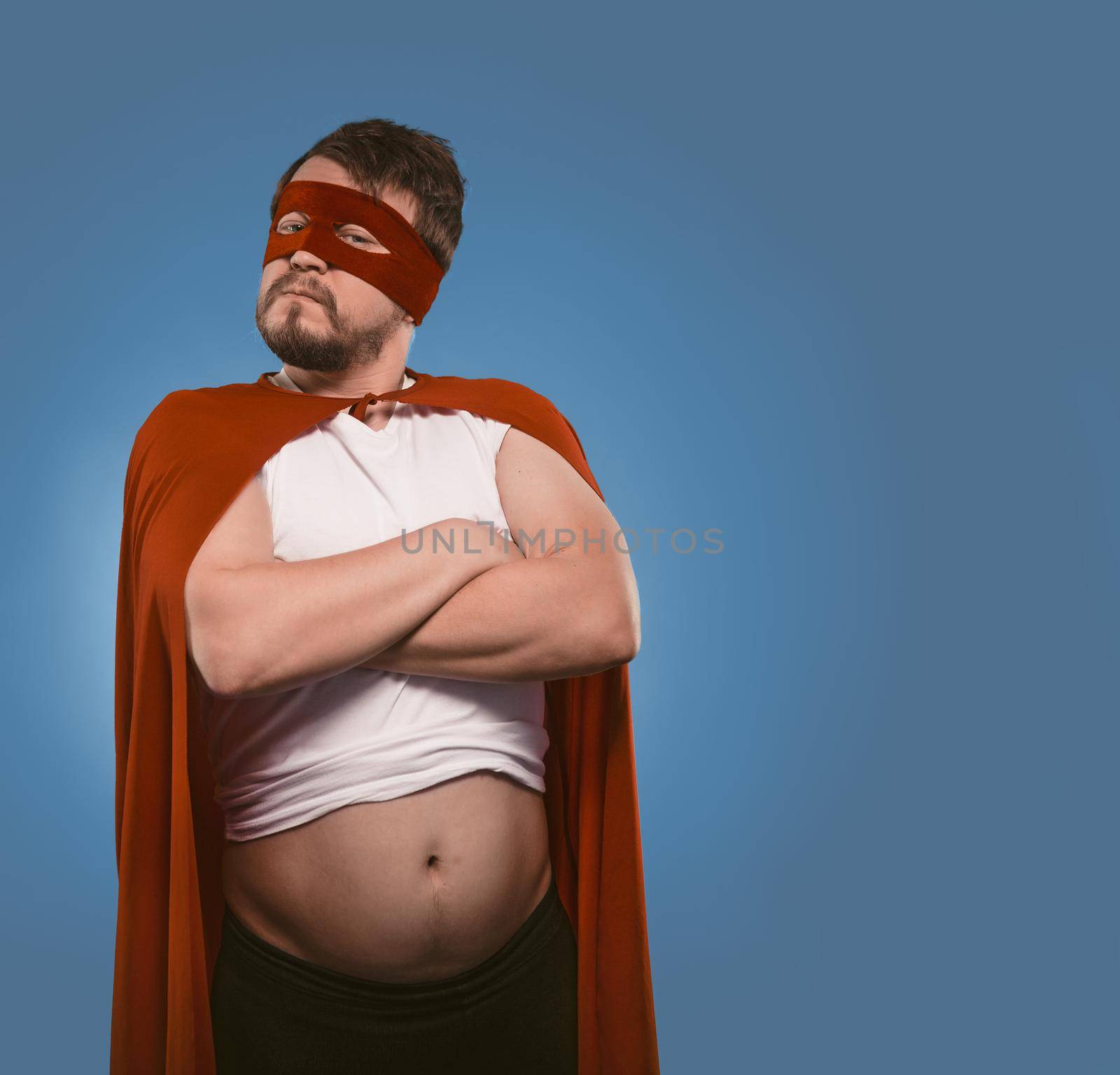 Serious super hero man posing with his arms crossed Confident bearded man in red superhero costume and mask on faded denim blue background. Copy Space on the right side by LipikStockMedia