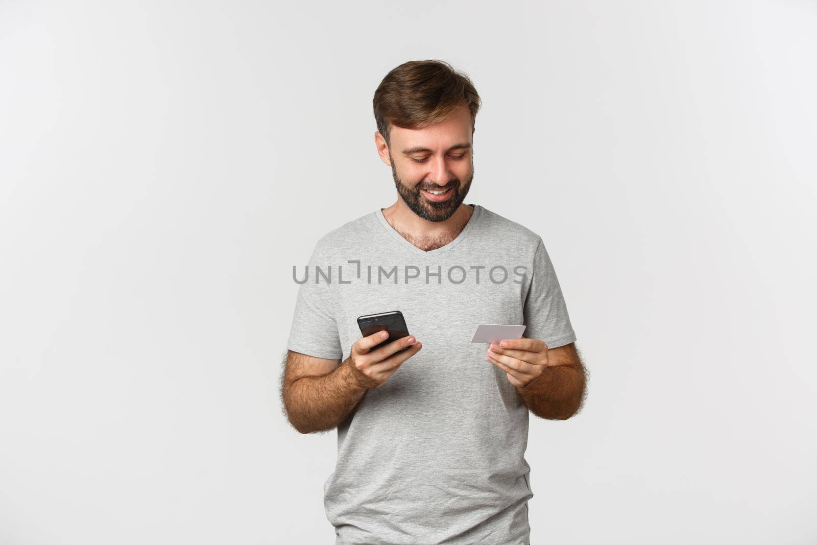 Handsome young man shopping online, holding credit card and mobile phone, standing over white background.