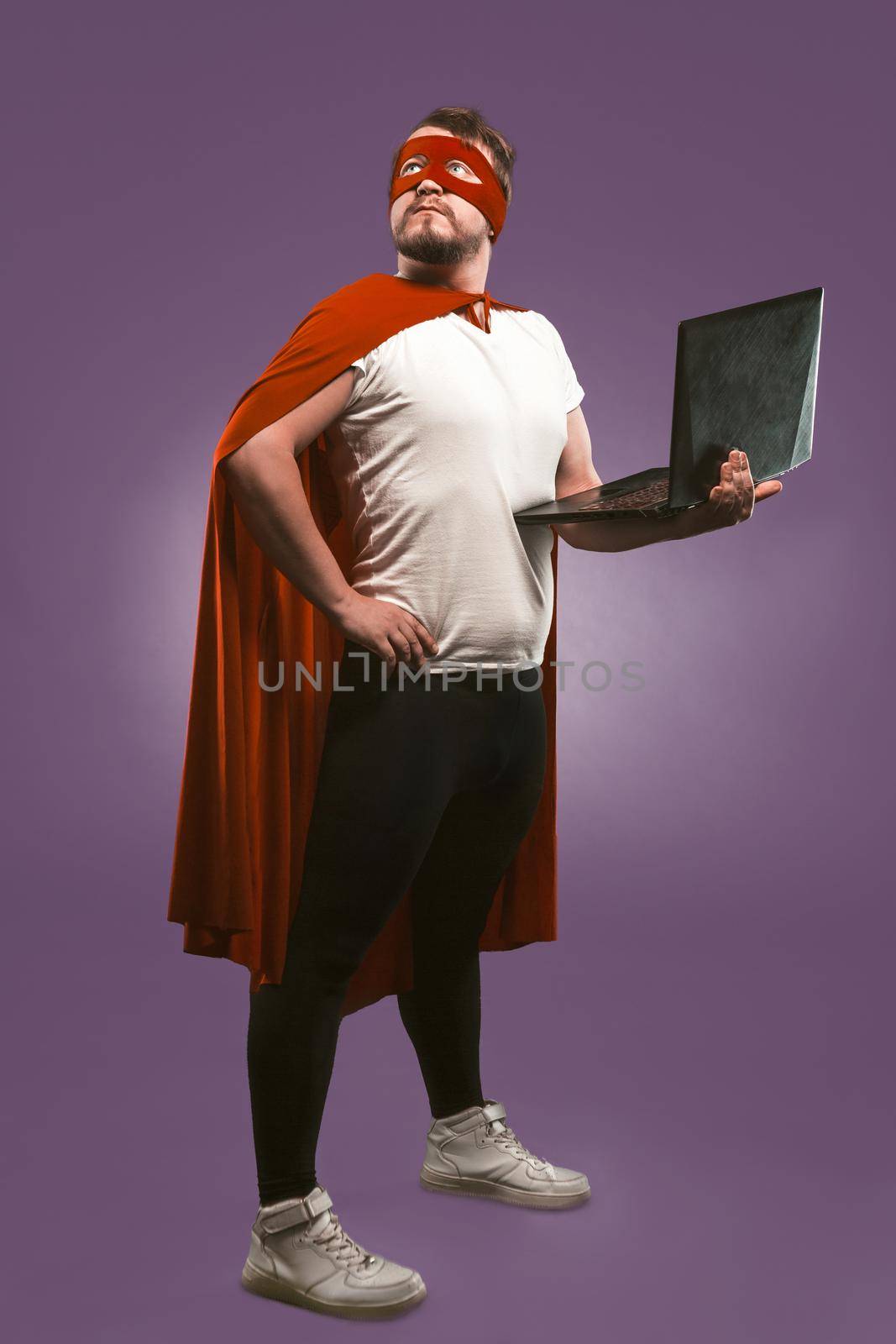 Super hero man holds a laptop looking up thoughtfully. Business man in red superhero costume using computer posing on grape purple background. Fast internet concept by LipikStockMedia