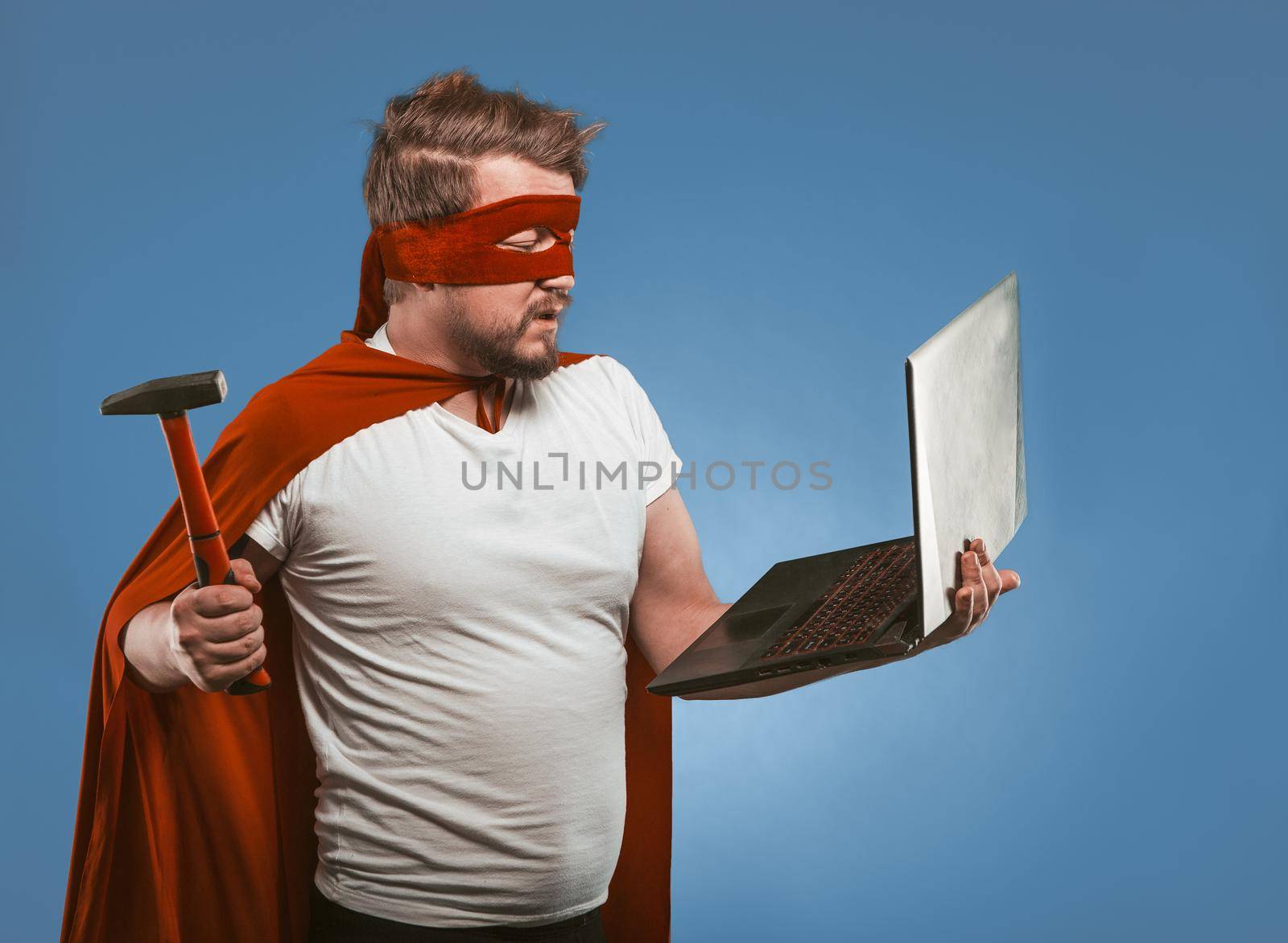 Super hero man going to smash or repair laptop computer with hammer. Side view of masked man in red super hero uniform posing on denim blue background. Computer hacking concept by LipikStockMedia