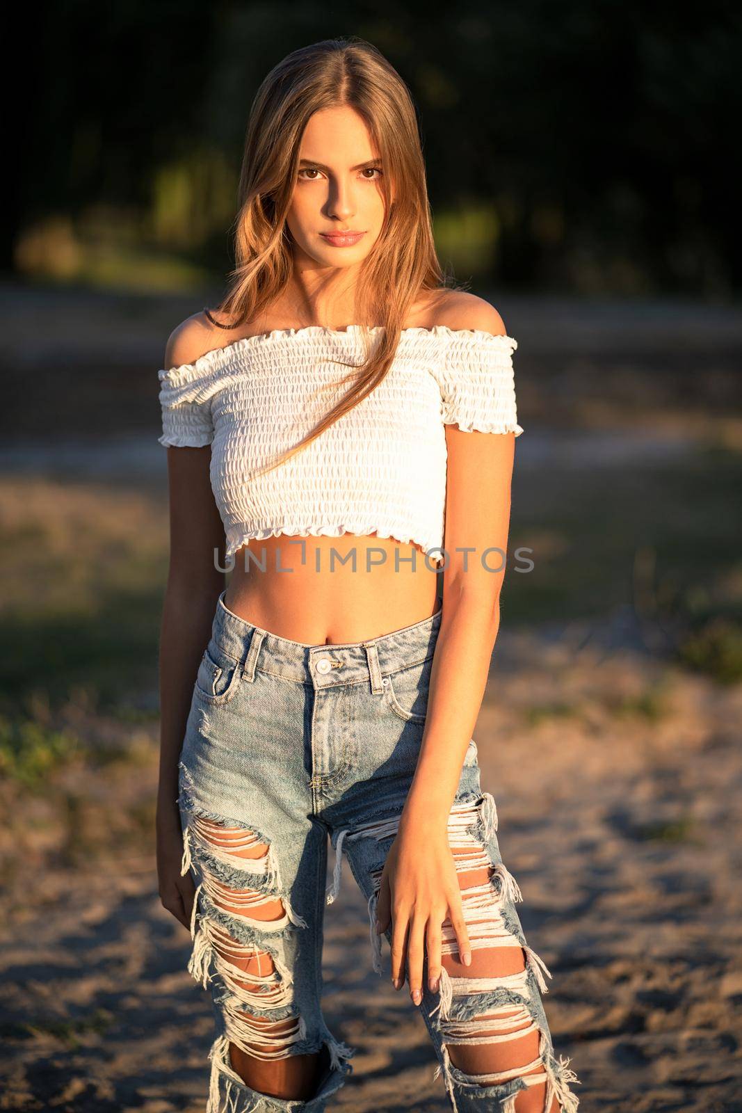 Hipster teenage girl looks at camera while posing on sunset outdoors. Fashion young model walks at beach in sunny evening by LipikStockMedia