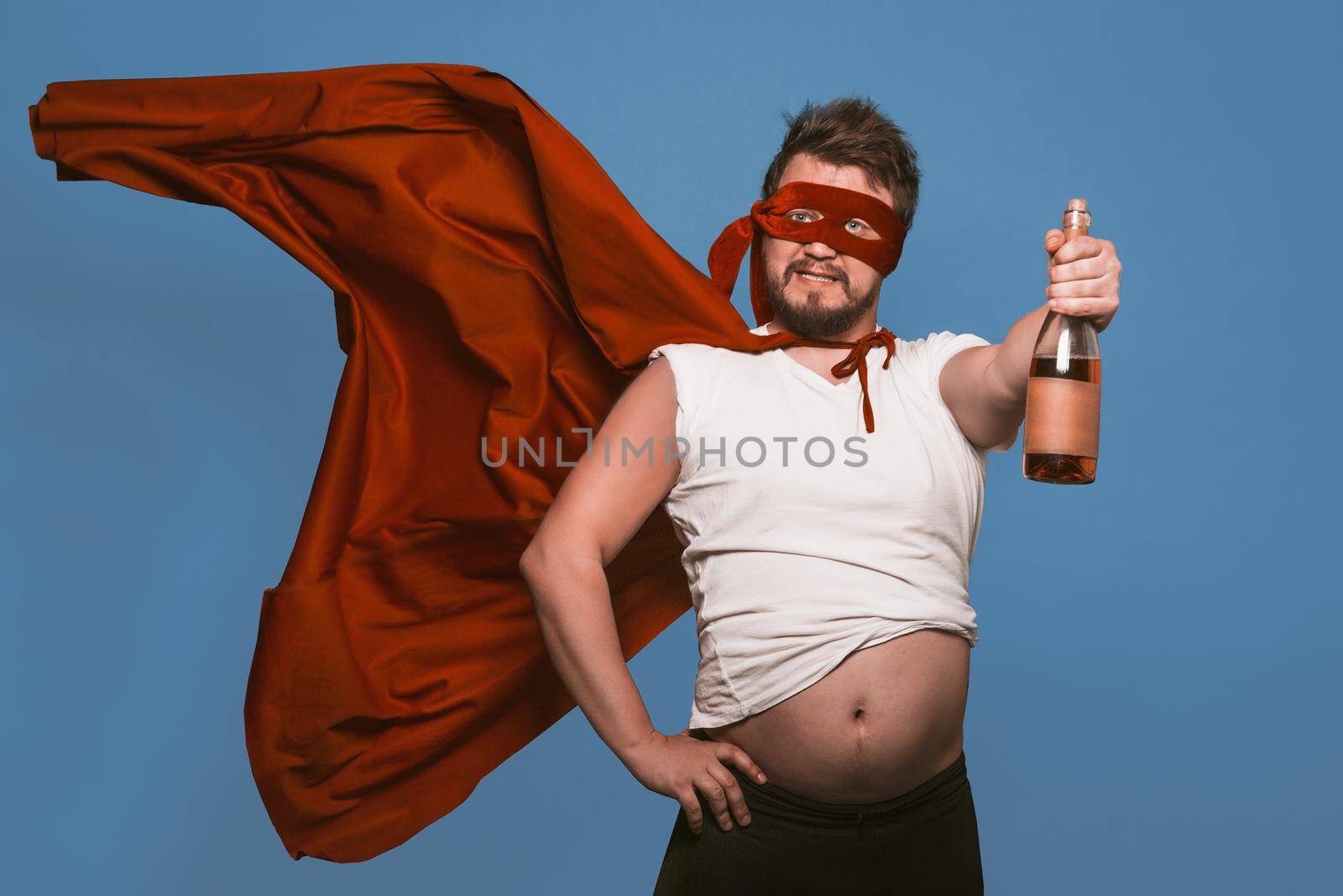 Tipsy superhero or antihero holds bottle of wine, man in superhero red mask with flying cloak holding bottle of alcohol while showing big tummy, isolated on faded denim blue background.