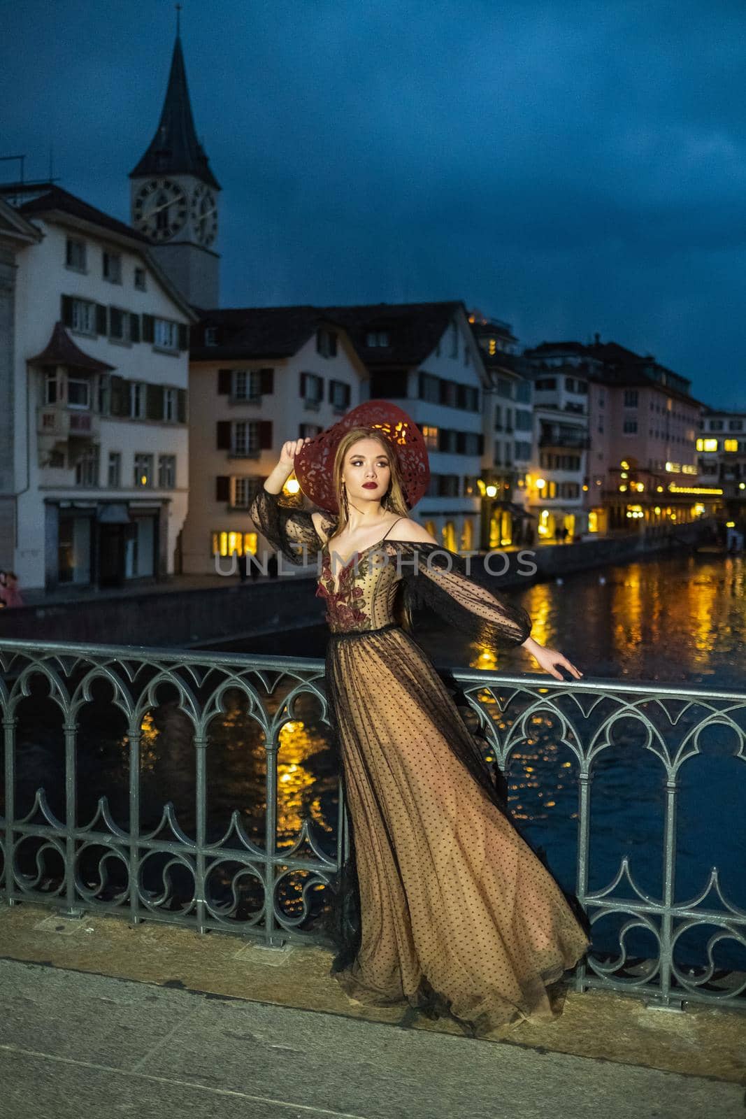 A stylish bride in a black wedding dress and a red hat poses at night in the old town of Zurich. Switzerland by Lobachad