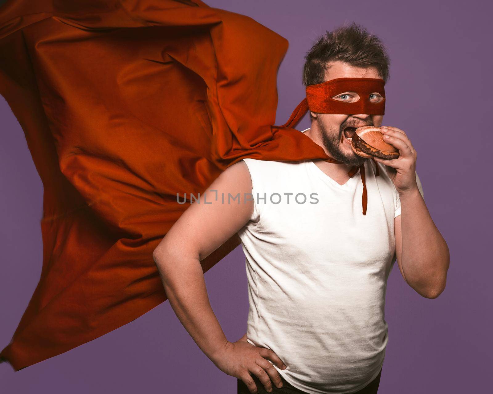 Hungry superhero man eats big hamburger with meat. Man in red flying cloak eats looking at camera on grape purple background. Fast food snack concept by LipikStockMedia