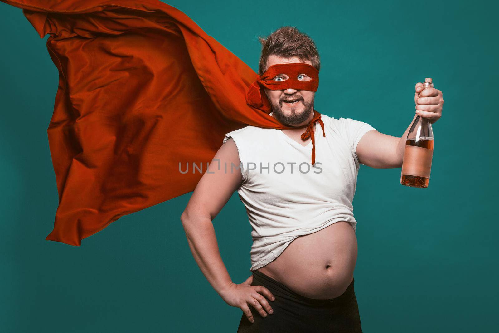 Drunk Superhero Or Antihero Man With Bottle Of Alcohol, Man In Superhero Red Mask And Fluttering Cloak Holding Bottle Of Wine Shows His Big Tummy, Isolated On Biscay Green Background by LipikStockMedia