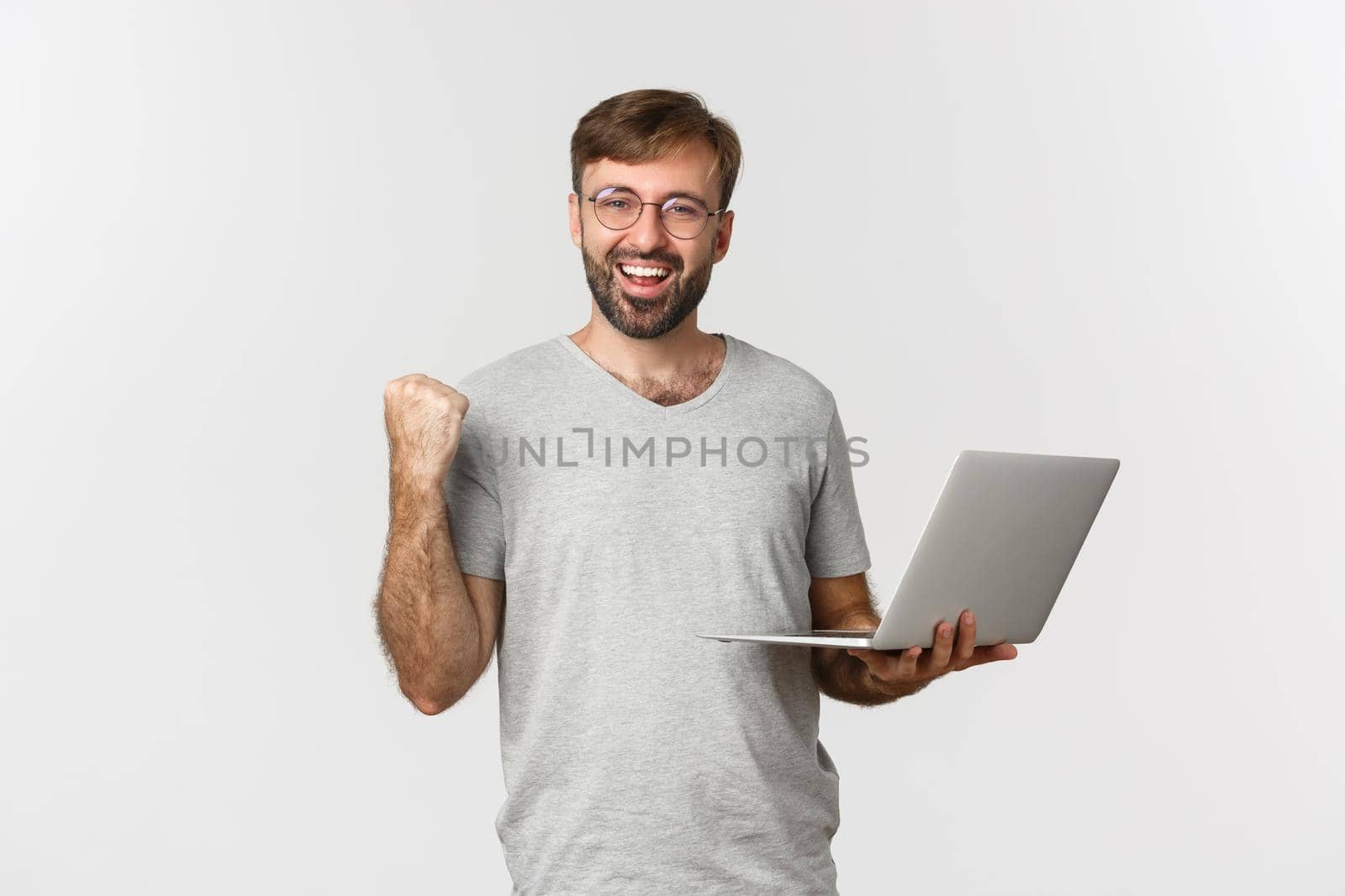 Portrait of cheerful handsome guy in glasses and gray t-shirt, holding laptop, achieve goal, winning something, standing over white background by Benzoix